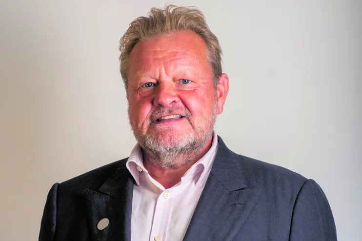 A middle-aged man with a beard and mustache, wearing a dark suit jacket and a white dress shirt, standing against a plain light background.
