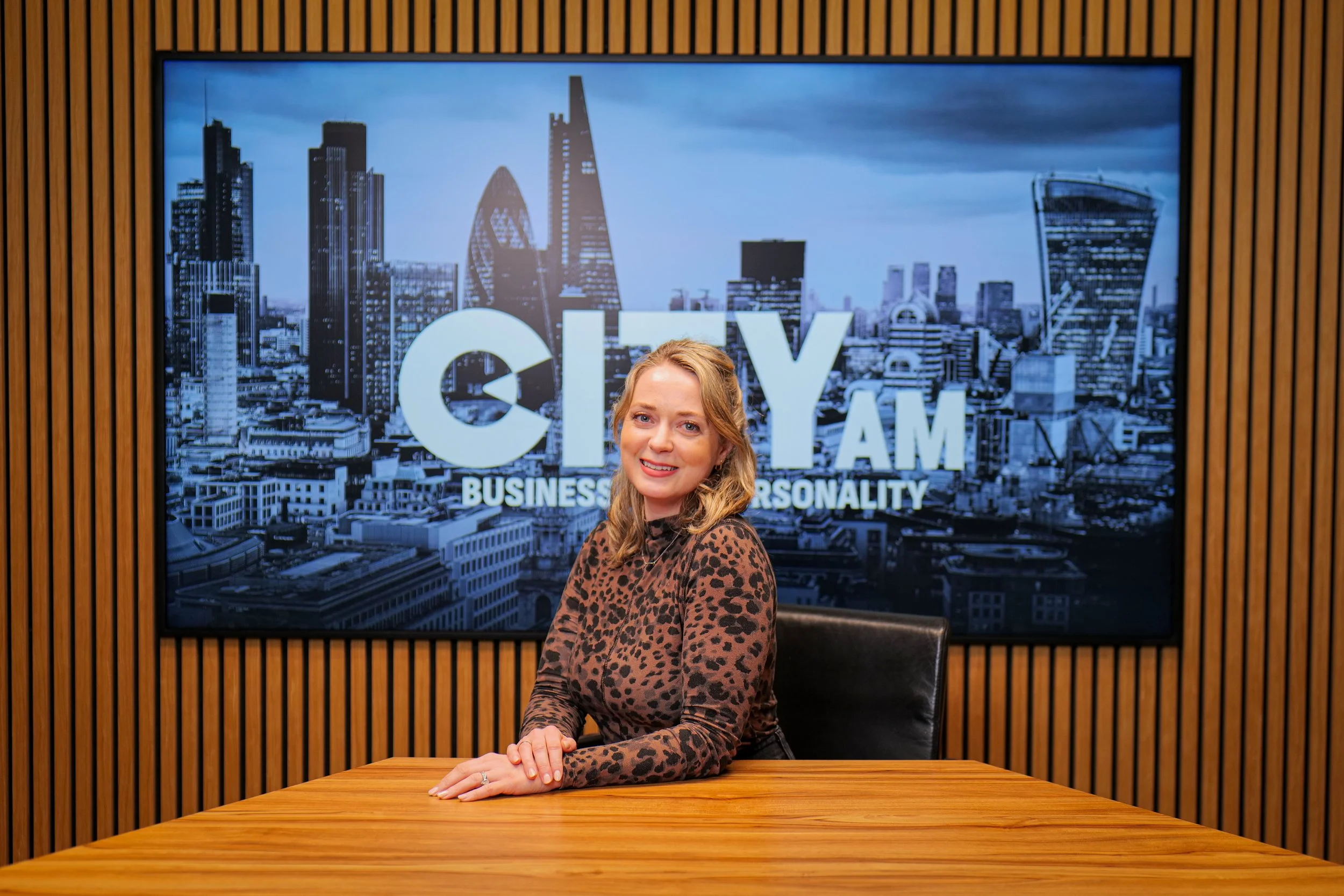A smiling woman with blonde hair wearing a gray sweater and dark blue jeans standing against a plain white background.