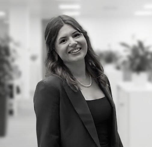 A middle-aged man with a beard and mustache, wearing a dark suit jacket and a white dress shirt, standing against a plain light background.