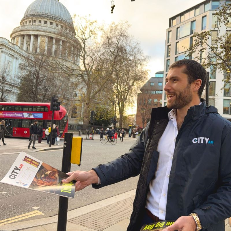 A man with a beard and short dark hair, wearing a CITY AM jacket, hands out a magazine on a city sidewalk with Us and St. Paul's Cathedral in the background.