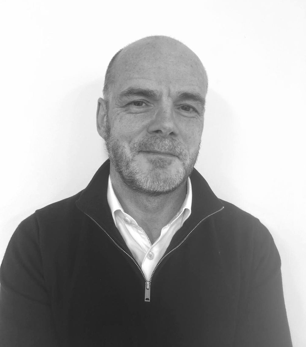 A black and white portrait of a middle-aged man with short hair, a beard, and a mustache, wearing a collared shirt and a zip-up jacket, standing against a plain white background.
