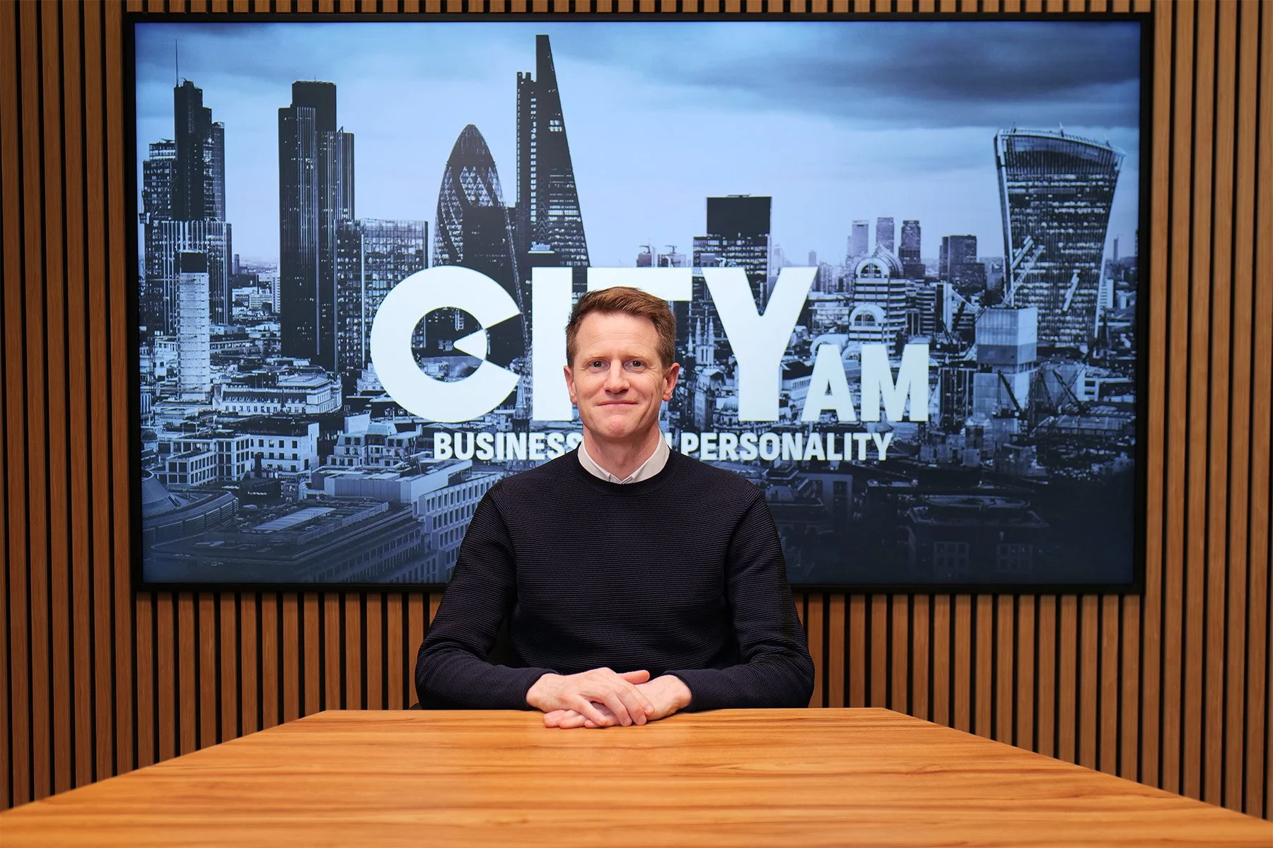 A middle-aged man in a black sweater sitting at a white table, facing the camera with a slight smile, against a plain light-colored background.