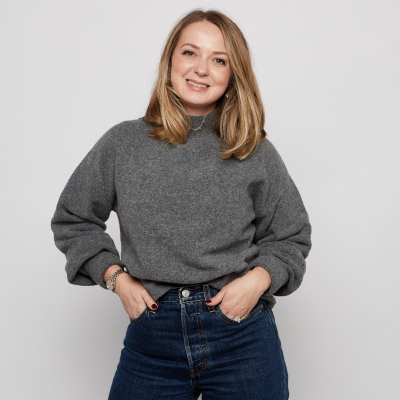 A smiling woman with blonde hair wearing a gray sweater and dark blue jeans standing against a plain white background.