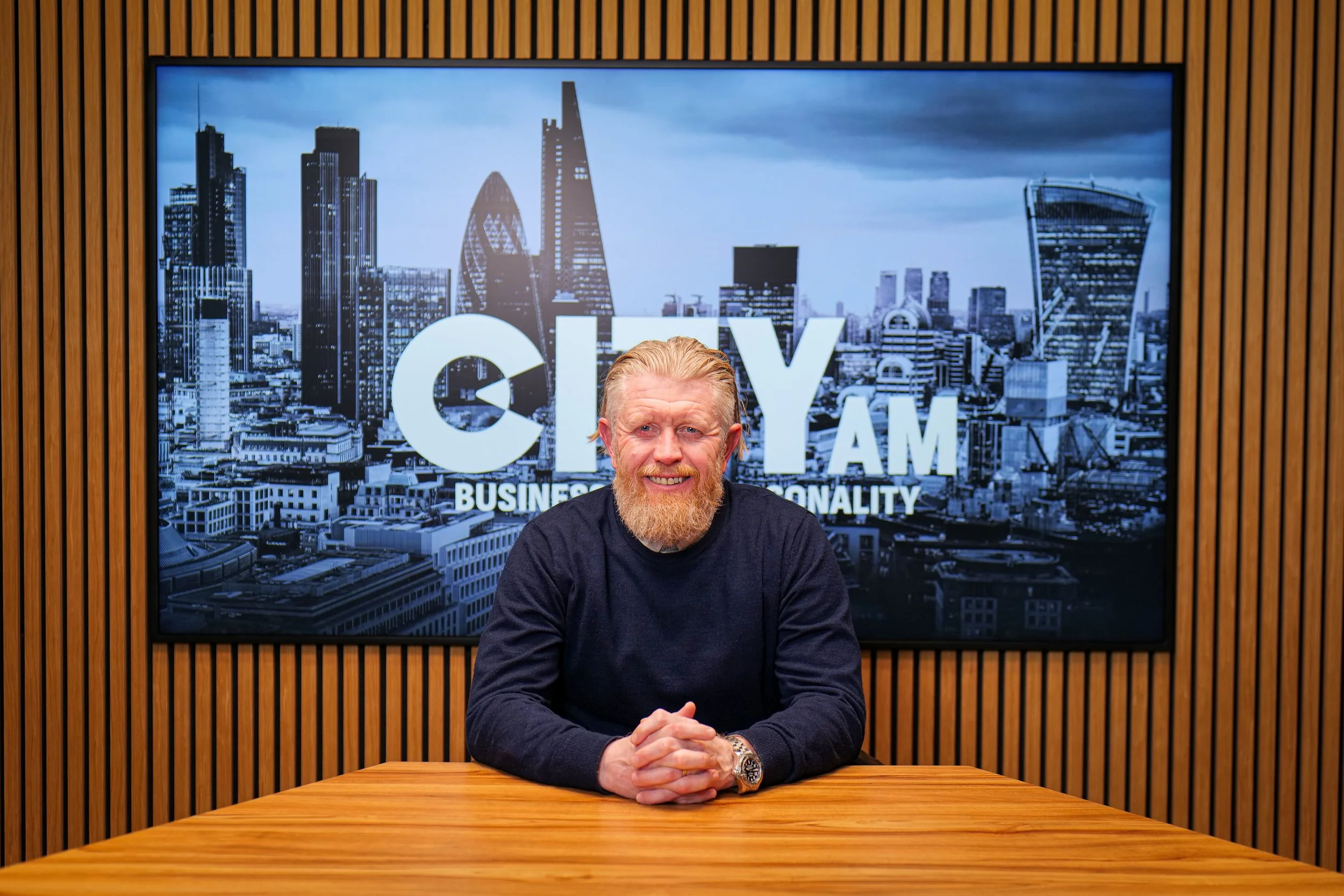 A smiling man with a beard and mustache, wearing a dark sweater over a collared shirt, in front of a plain white background.