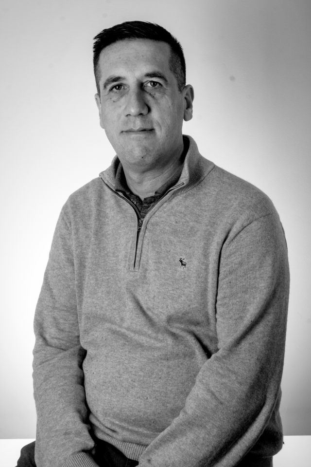 A man with short dark hair and light skin, sitting against a plain background, wearing a light-colored sweater with a zipper and a small logo on the chest.