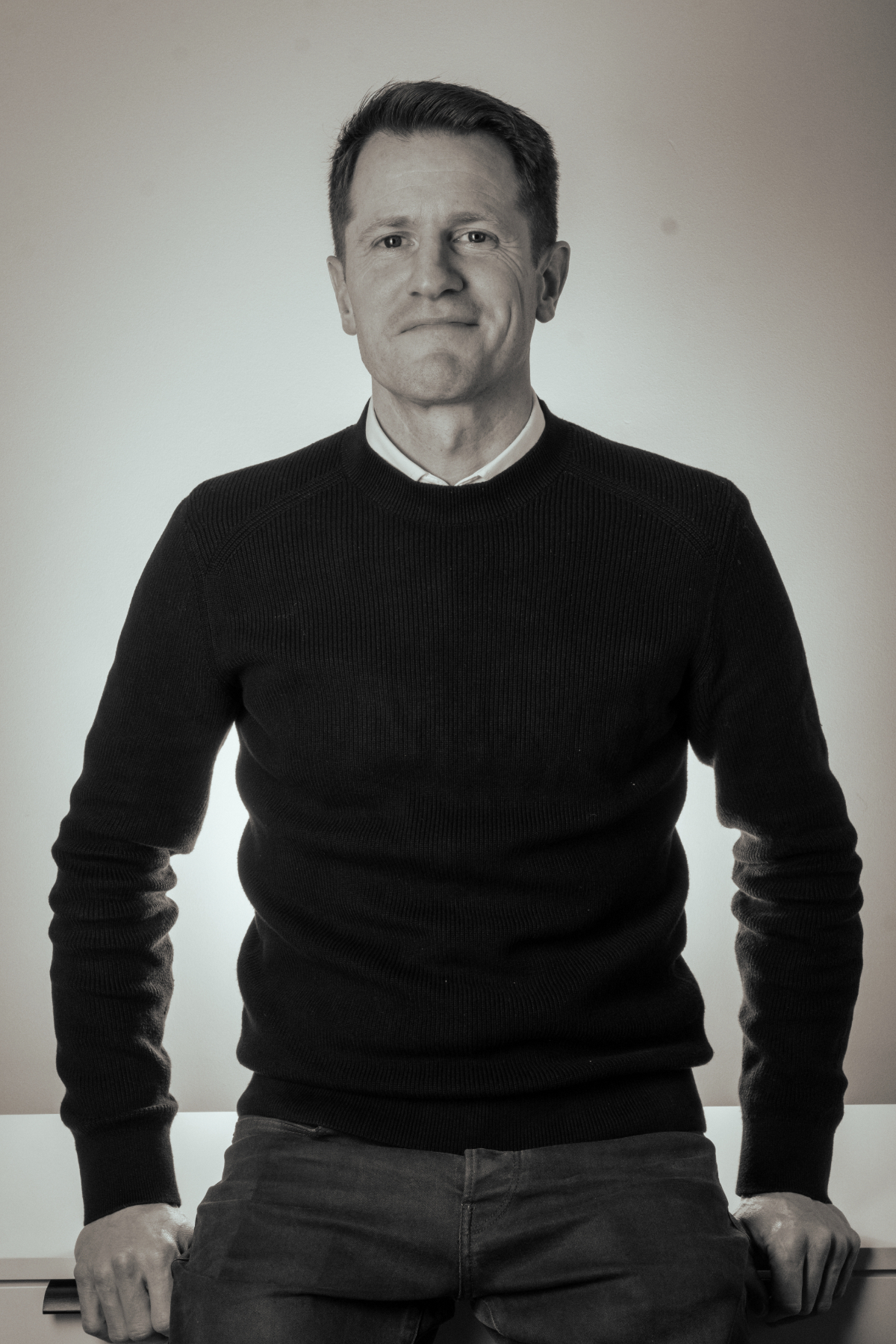 A middle-aged man in a black sweater sitting at a white table, facing the camera with a slight smile, against a plain light-colored background.