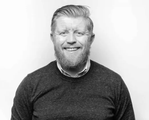 A smiling man with a beard and mustache, wearing a dark sweater over a collared shirt, in front of a plain white background.