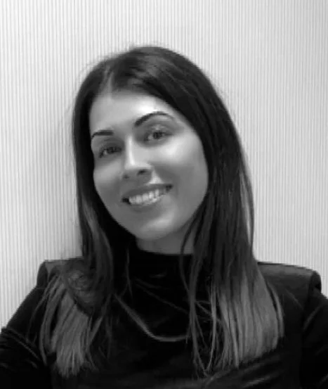 Black and white photo of a smiling woman with shoulder-length dark hair, wearing a dark top, standing against a textured wall.
