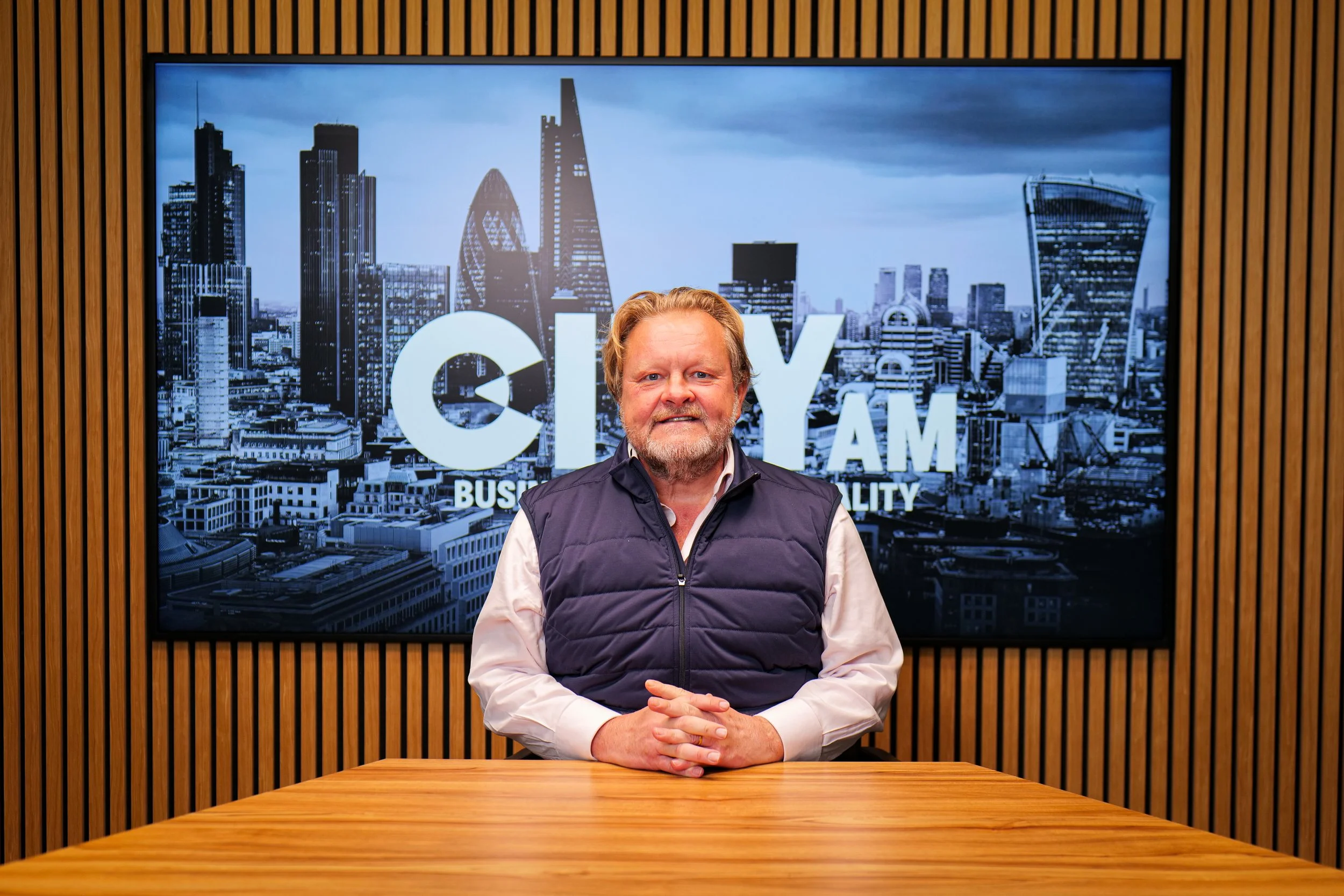 A middle-aged man with a beard and mustache, wearing a dark suit jacket and a white dress shirt, standing against a plain light background.