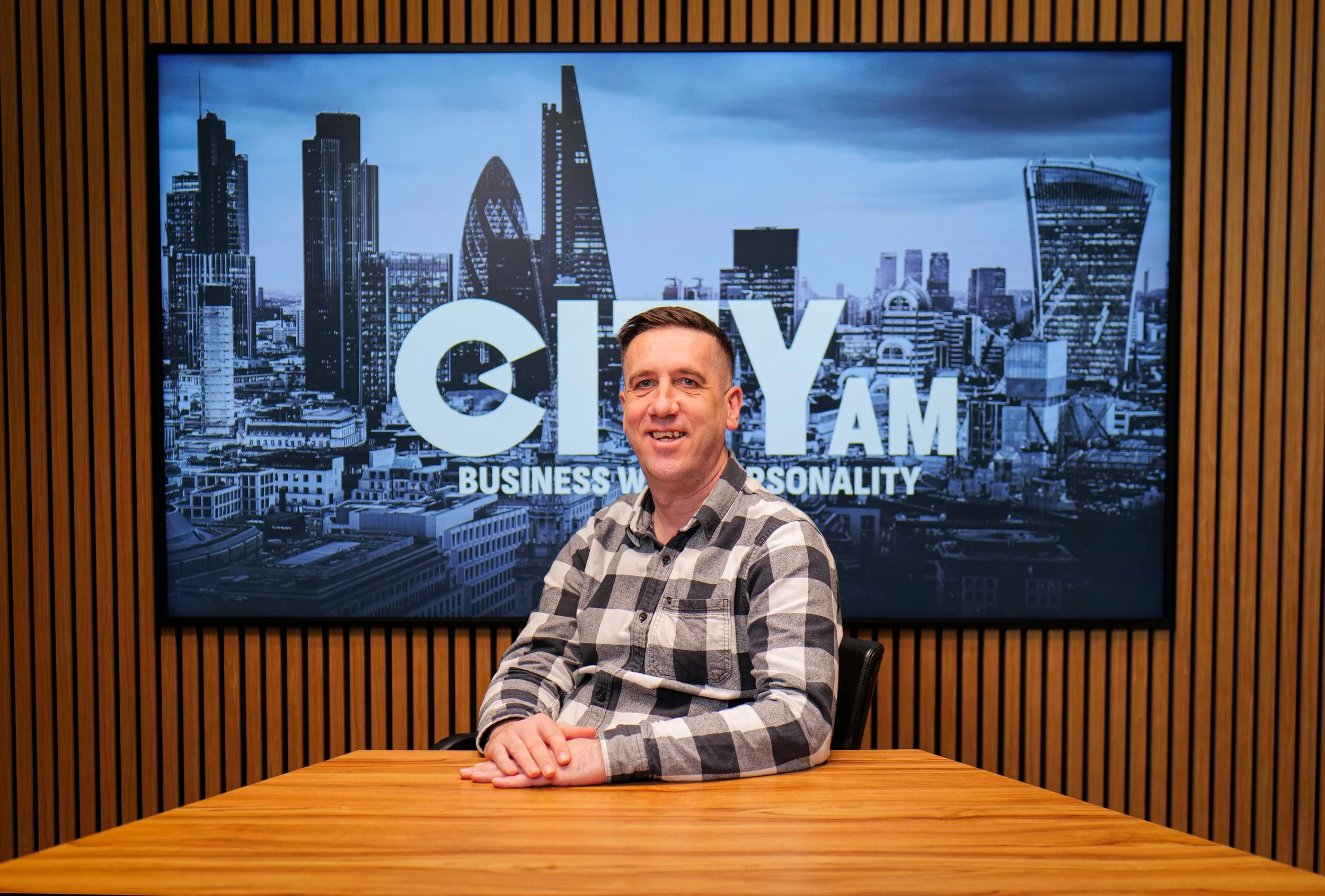 A man with short dark hair and light skin, sitting against a plain background, wearing a light-colored sweater with a zipper and a small logo on the chest.