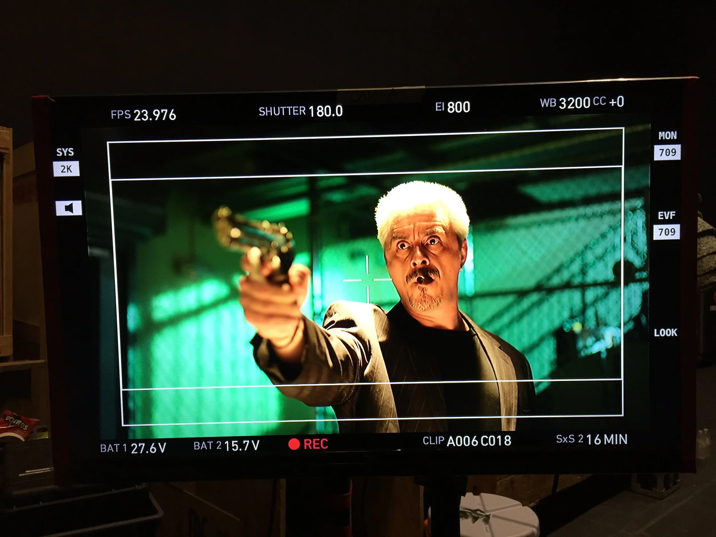 A scene from a film or TV show displayed on a monitor shows a man with white hair pointing a gun at the camera with a serious expression, in an industrial setting with green lighting.
