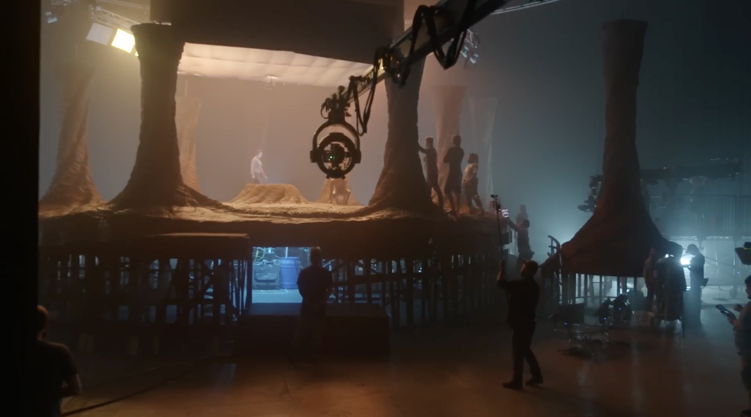 Behind the scenes of a film or TV show set, with a large suspended platform resembling an alien planet landscape, featuring large tree-like structures and a sandy ground, crew members, and equipment working in a dimly lit environment.