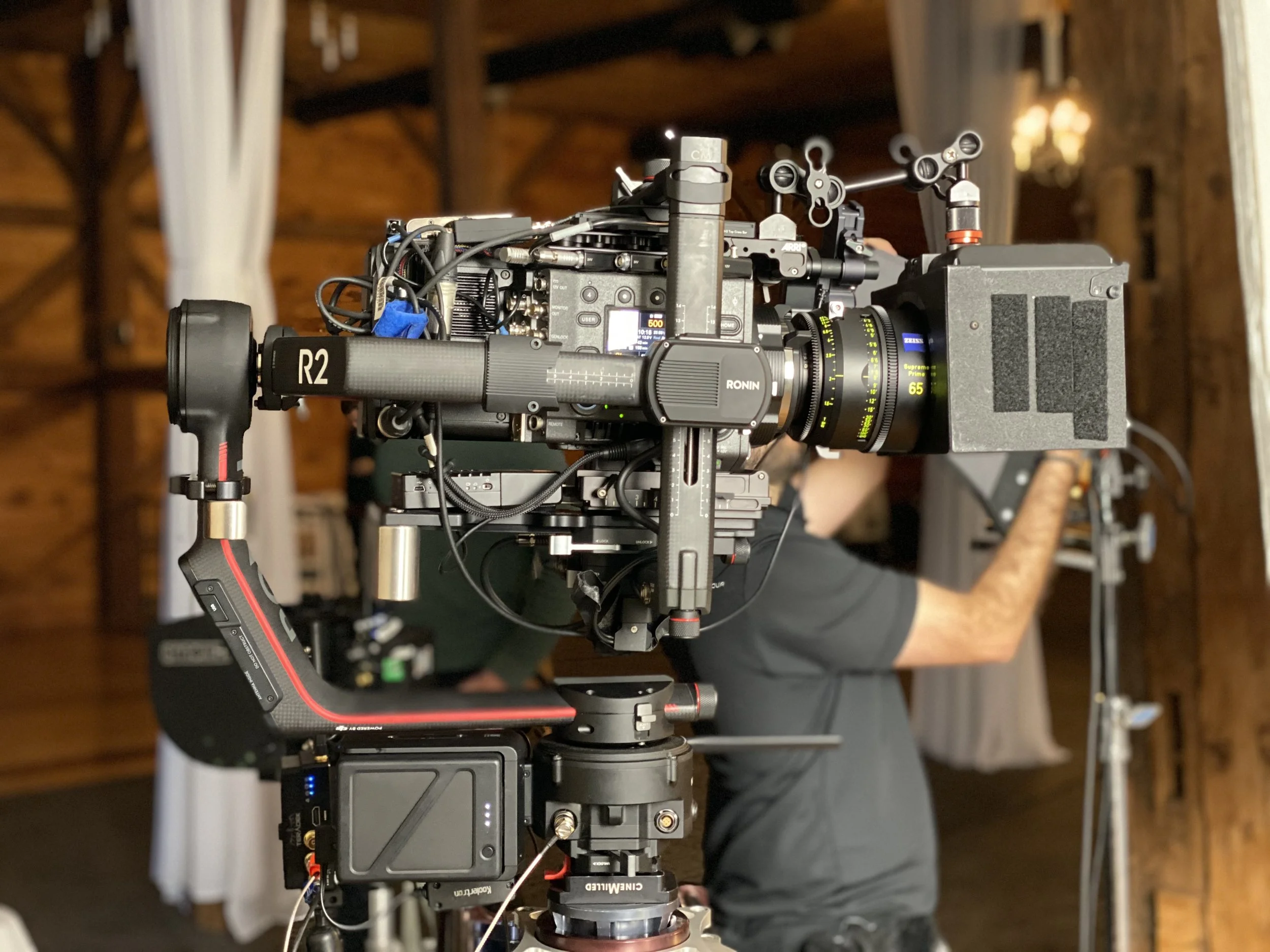 Close-up of a high-end professional film camera mounted on a tripod in a wooden studio. The camera is equipped with various accessories, lenses, and electronic controls. A crew member wearing a dark shirt is seen adjusting equipment in the background.