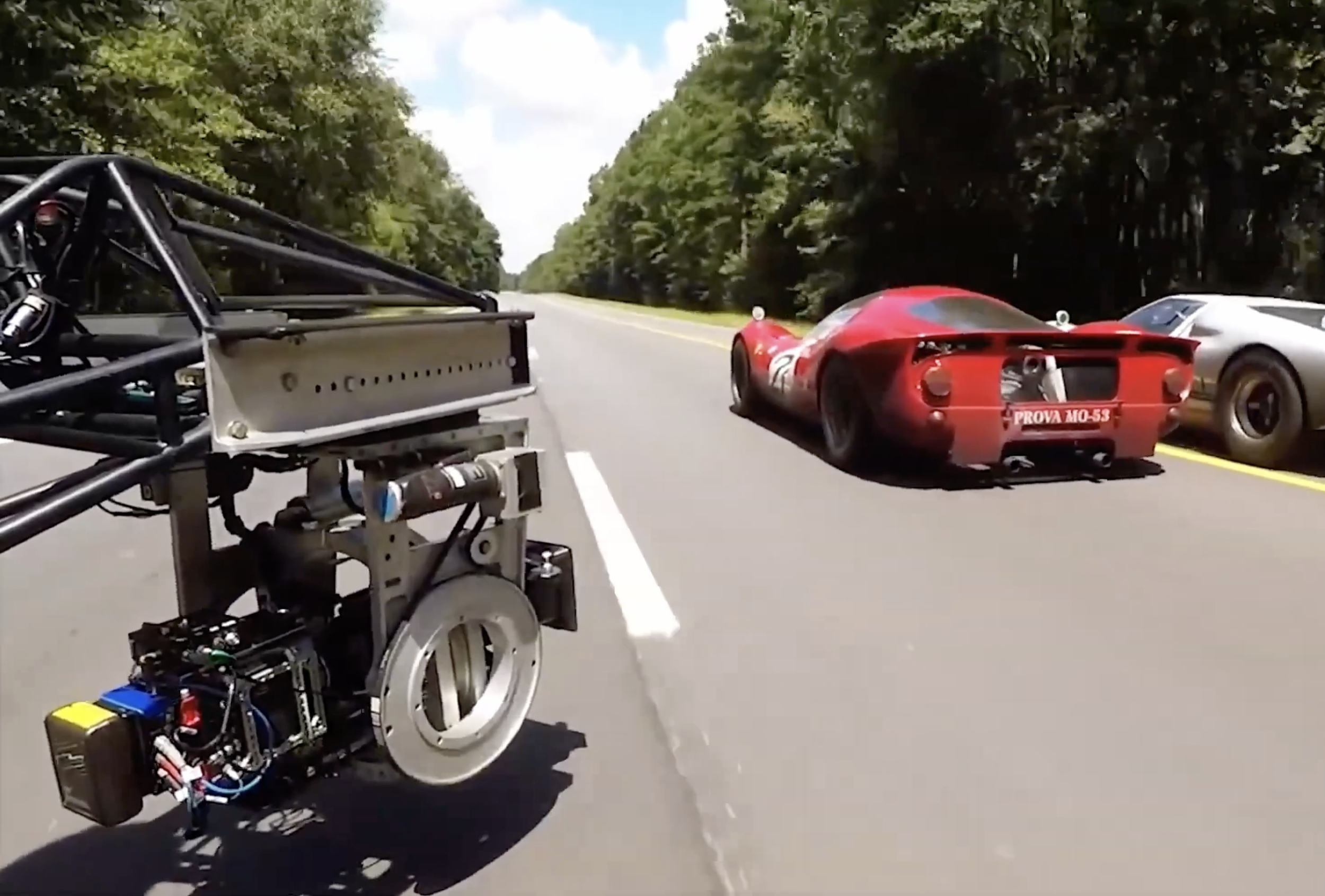 A red vintage race car on a two-lane road surrounded by trees, with a modern silver car beside it and a robotic device or drone in the foreground.