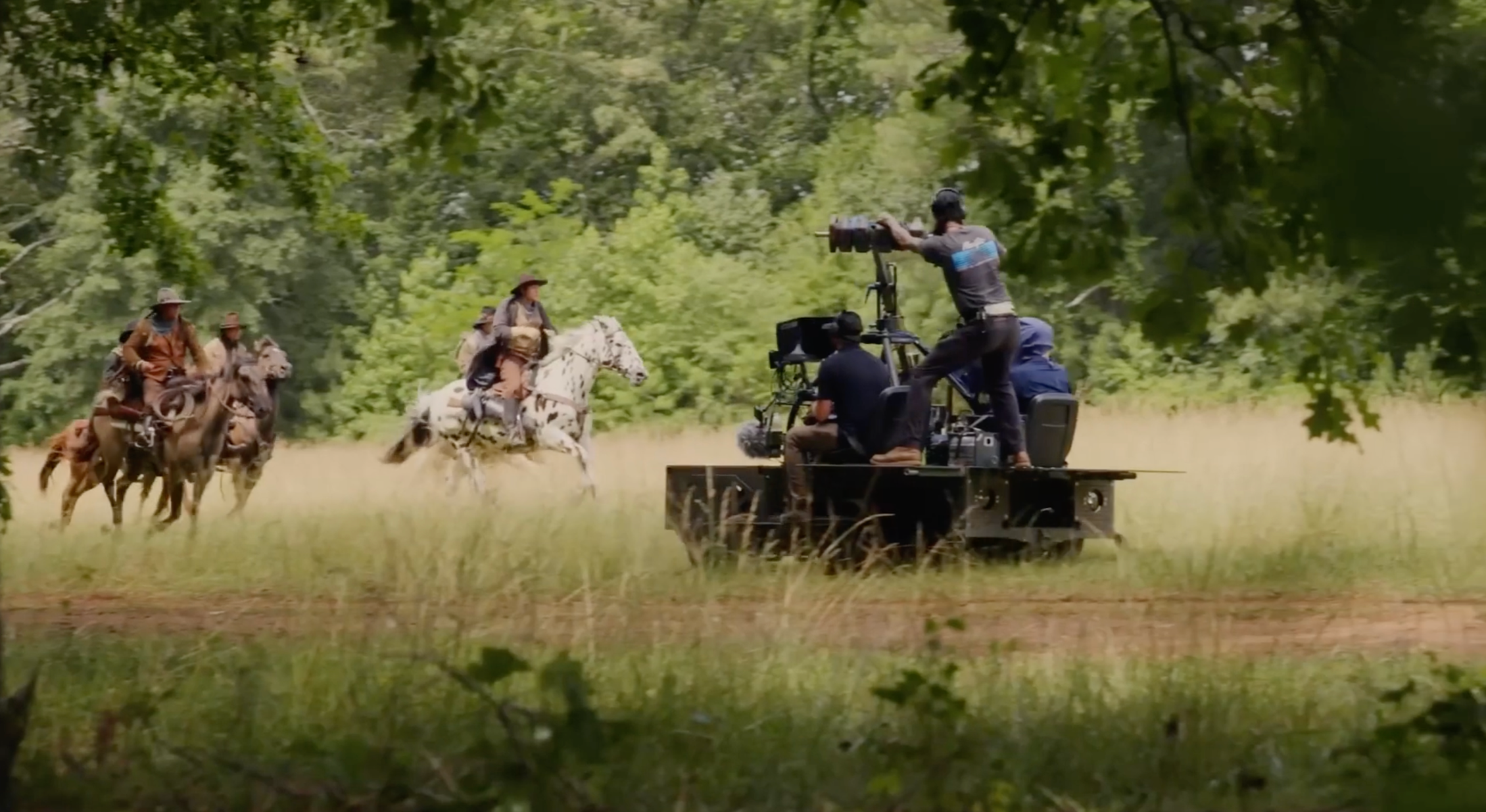 A film crew filming a scene in a wooded outdoor setting with three people on horseback riding through tall grass, and a camera operator on a camera dolly capturing the scene.
