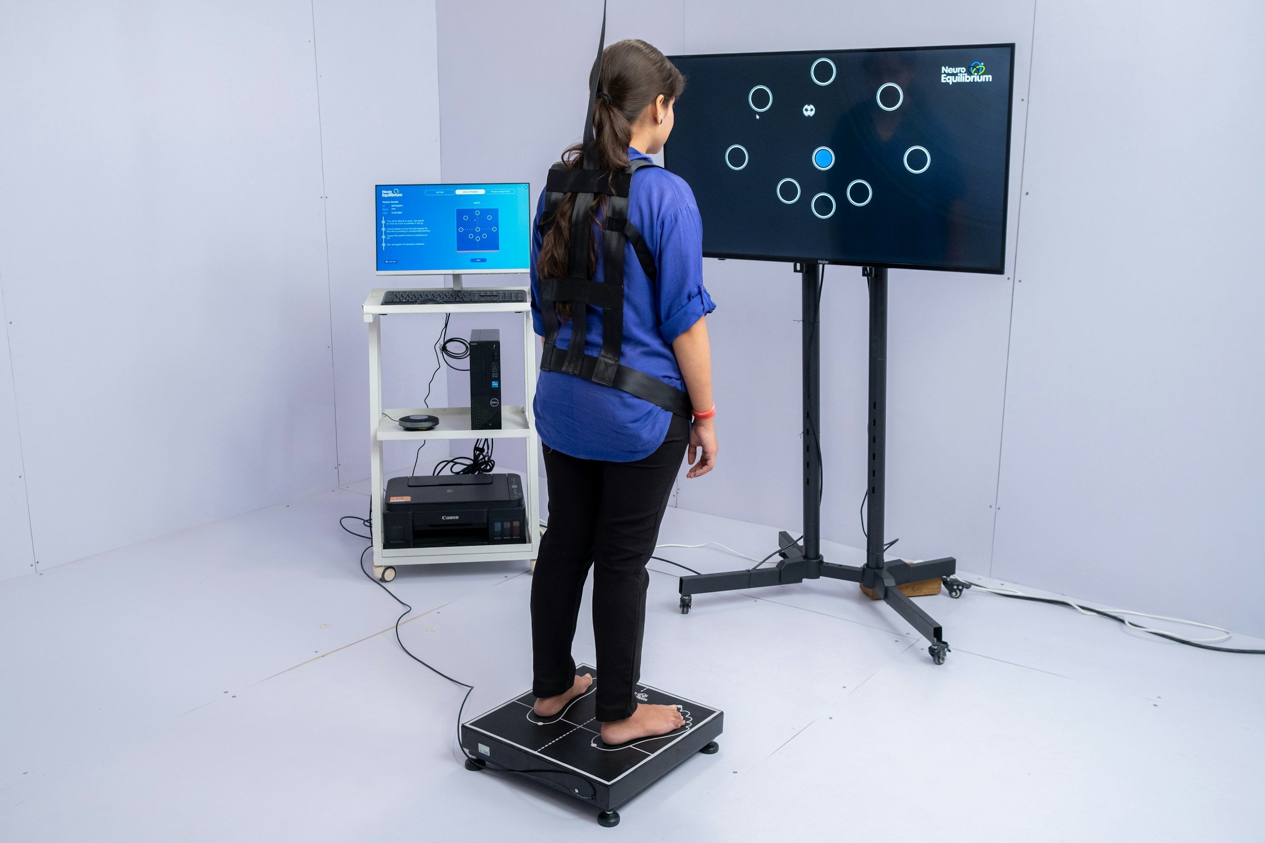 A woman standing on a pressure-sensitive platform connected to a small monitor and larger display screen, participating in a neurofeedback or balance test in a clinical or research setting.