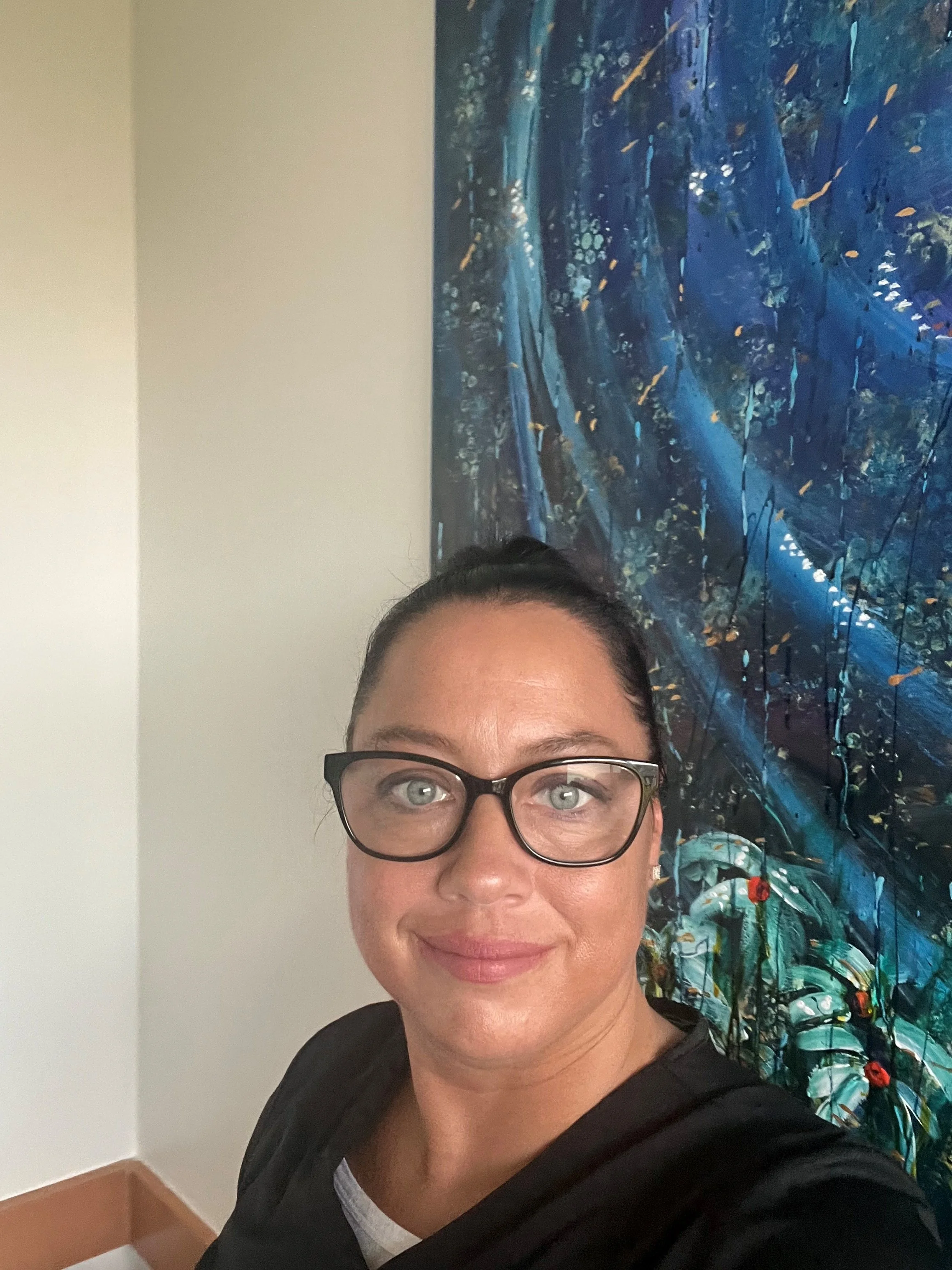 A woman with glasses and dark hair taking a selfie in front of a colorful abstract painting in a gallery or museum.