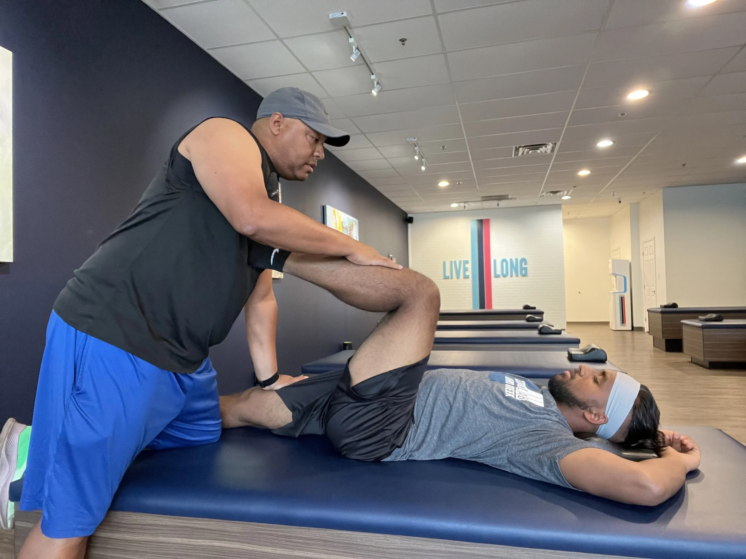 A person in a black tank top and gray cap providing physical therapy to a man lying face down on a massage table, with his arm extended and hand resting on his back, in a fitness or therapy center.
