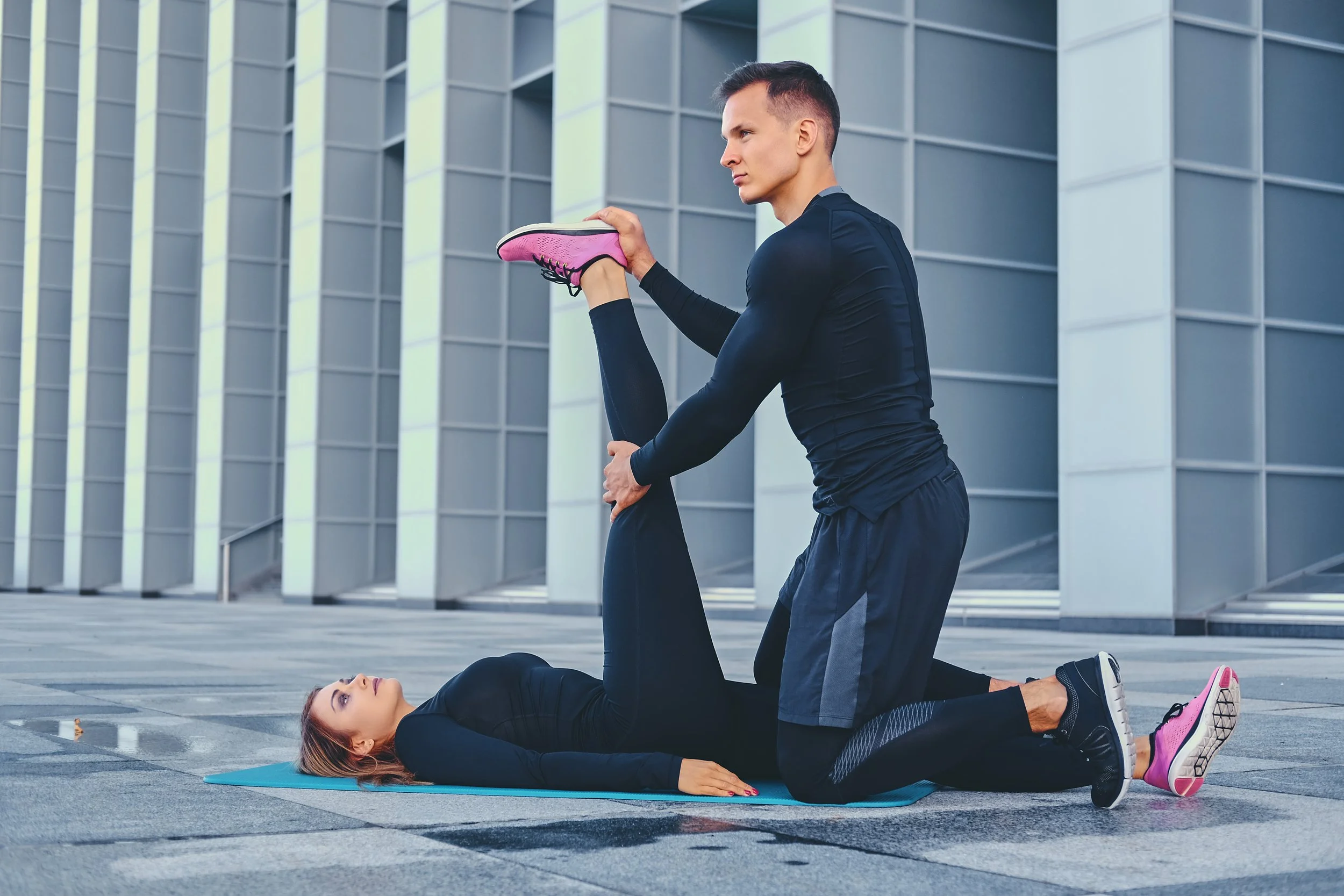 A man assisting a woman with stretching exercises on a yoga mat outdoors in front of a modern building.