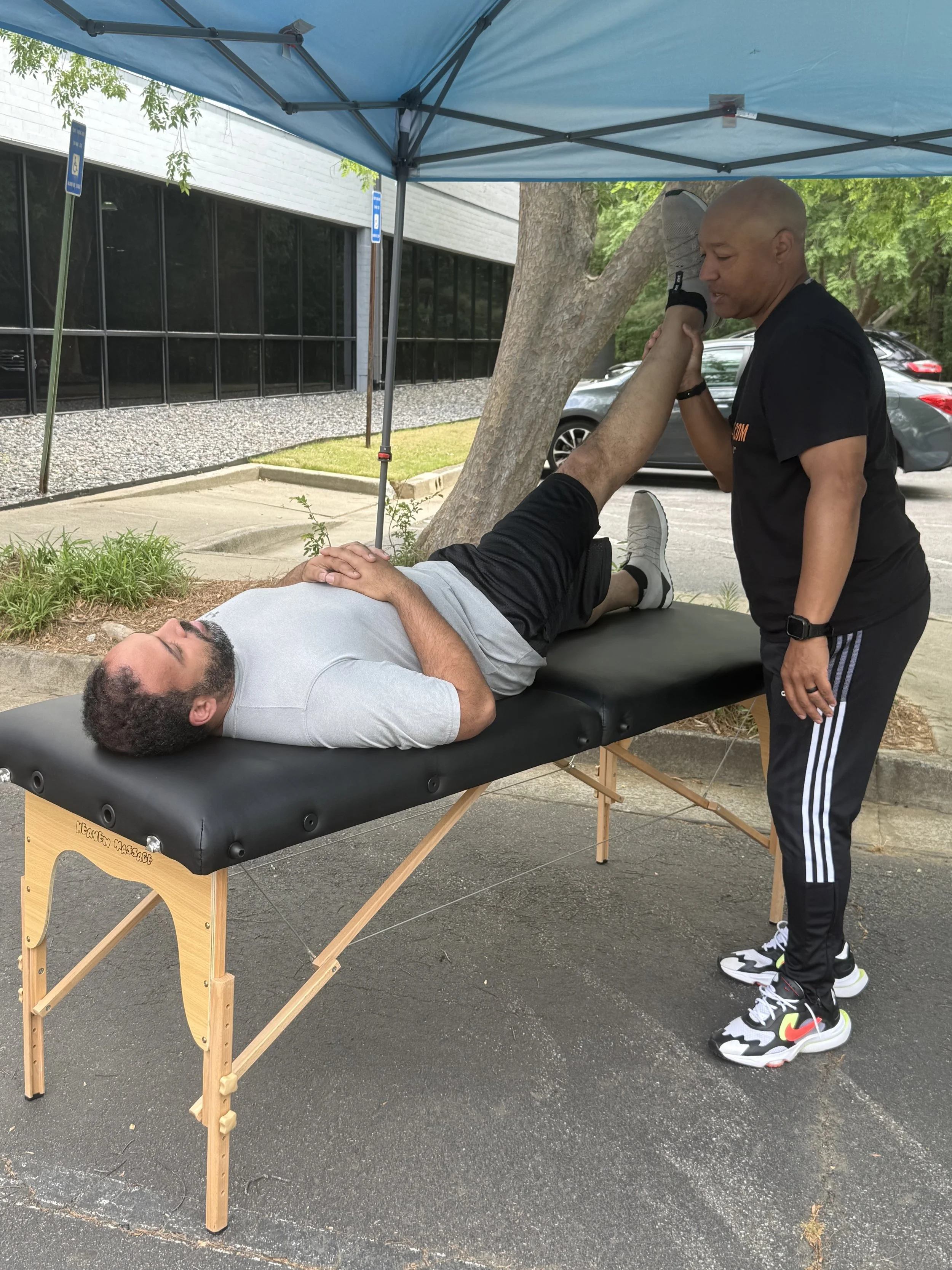 A massage therapist is stretching a man's leg during an outdoor massage session under a blue canopy, with parked cars and a building in the background.