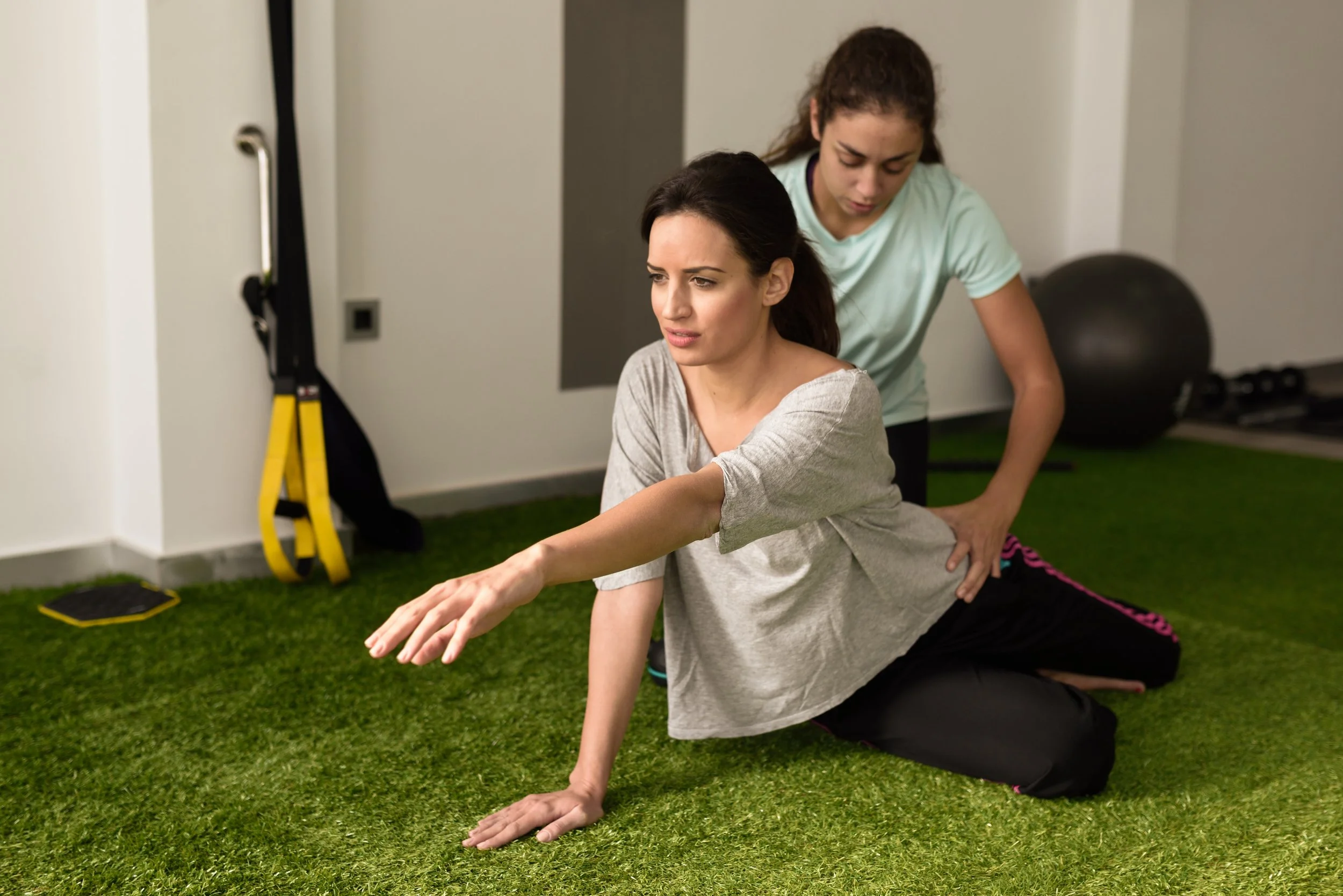 A woman is performing a stretching exercise on a green gym mat while a trainer assists her from behind in a gym.