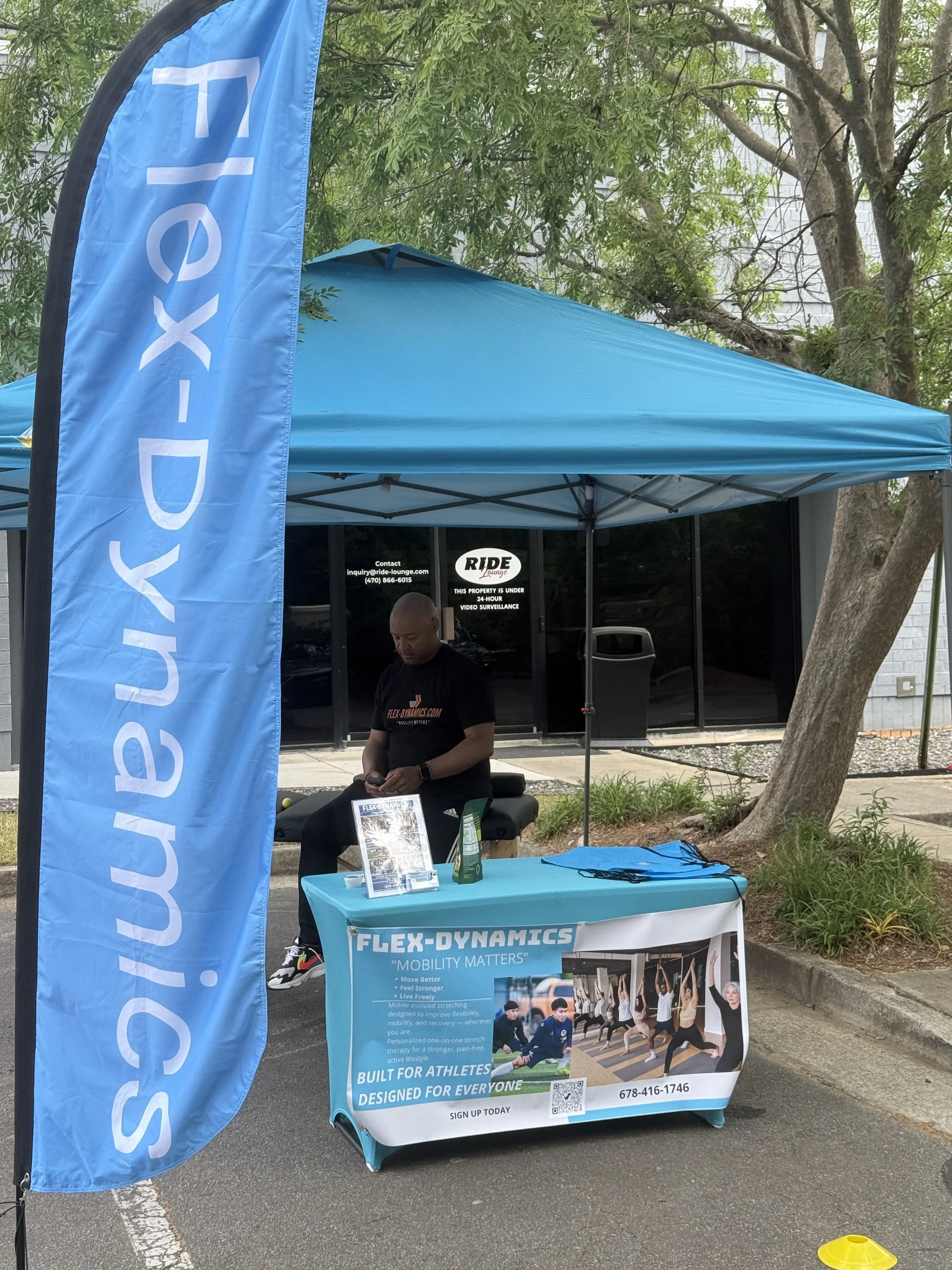 Outdoor booth with a blue canopy tent and a blue banner that reads 'SIX' in large letters. A man is standing behind a table with promotional material and a poster visual showing group exercise activities. The setting includes trees and a building in the background, and the scene appears to be at a community event or health fair.