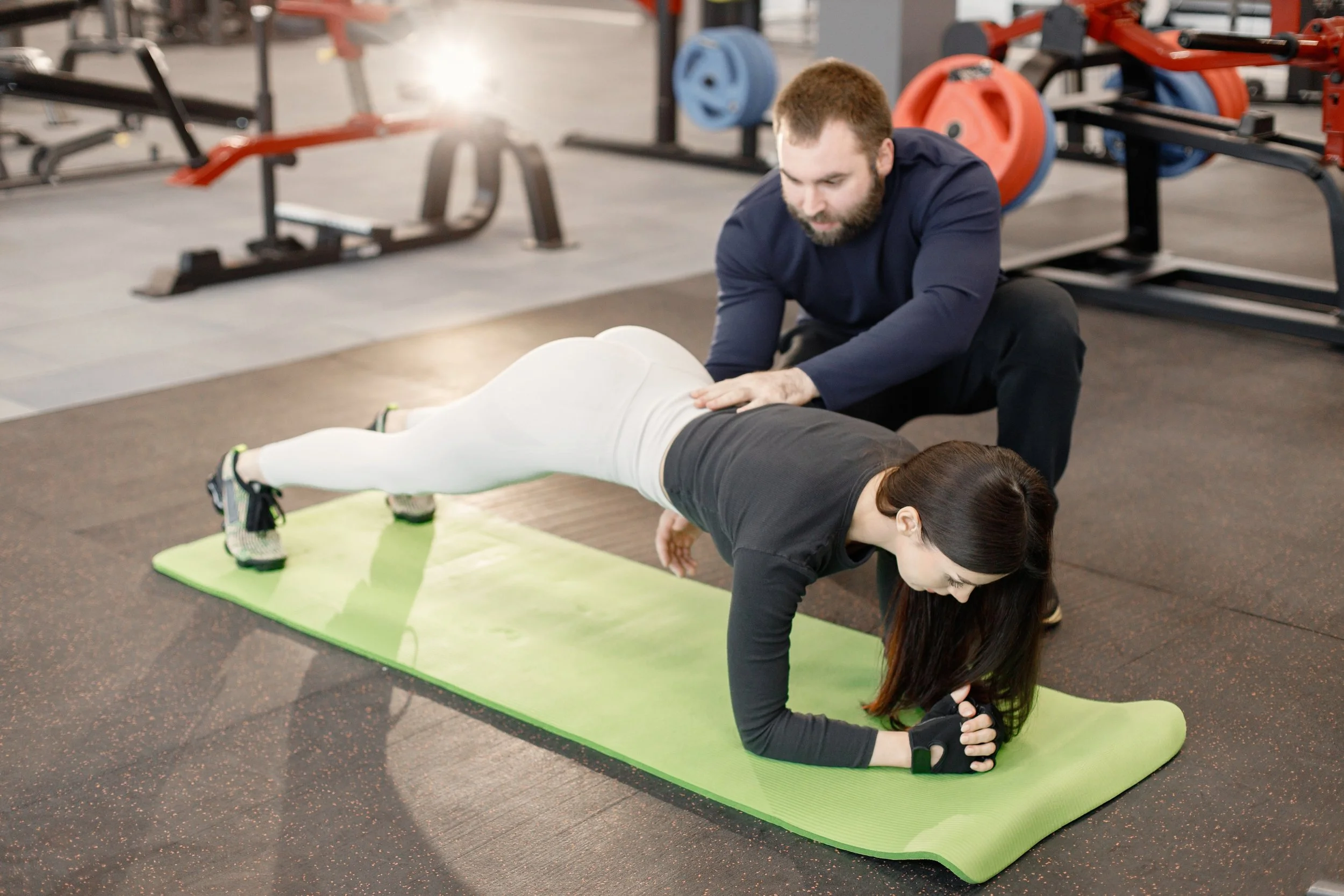 A woman is doing a plank exercise on a green yoga mat in a gym, with a trainer assisting her.