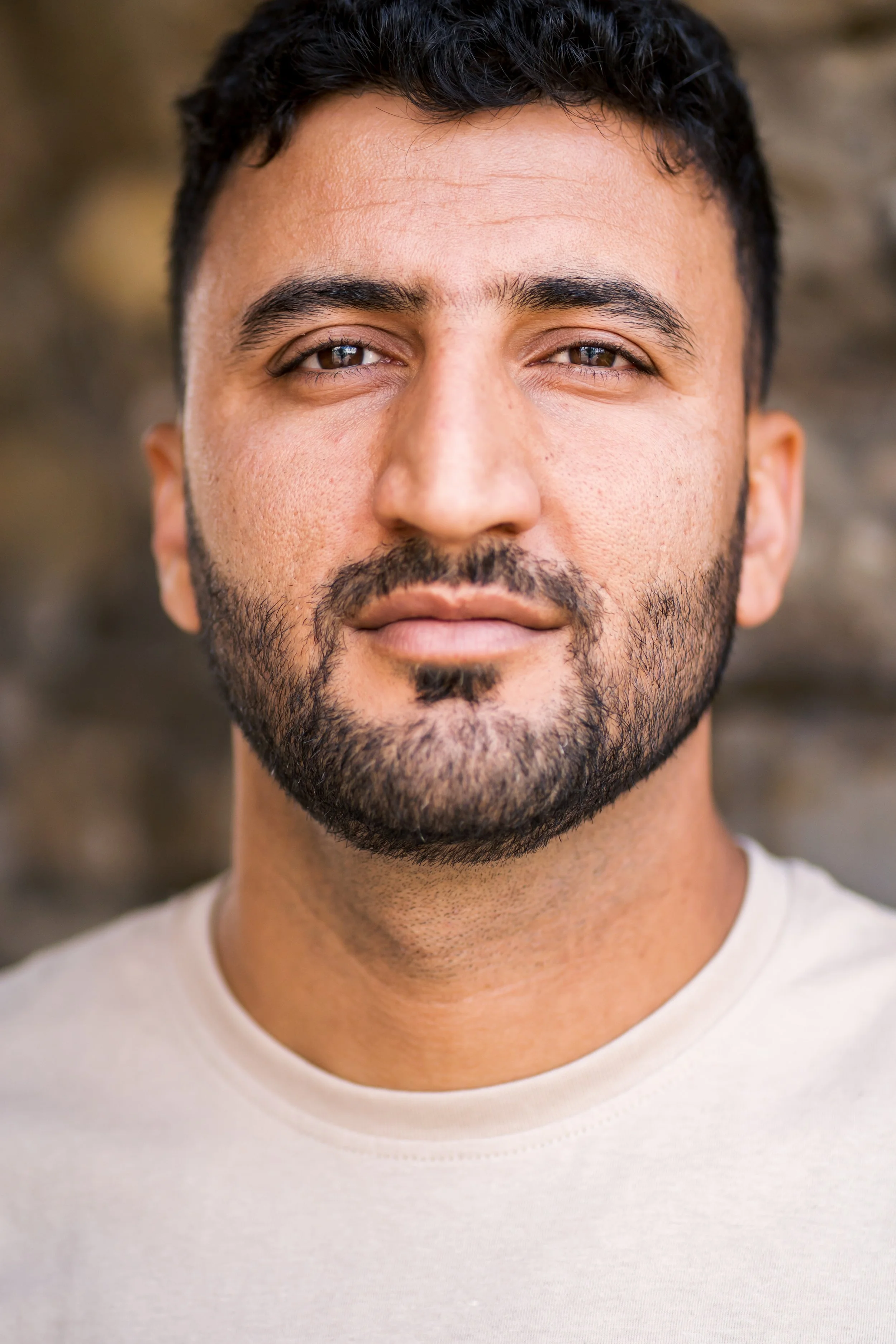 Nahaufnahme eines Mannes mit dunklem Haar, Bart und braunen Augen, trägt ein helles T-Shirt, im Hintergrund unscharfer Naturhintergrund.