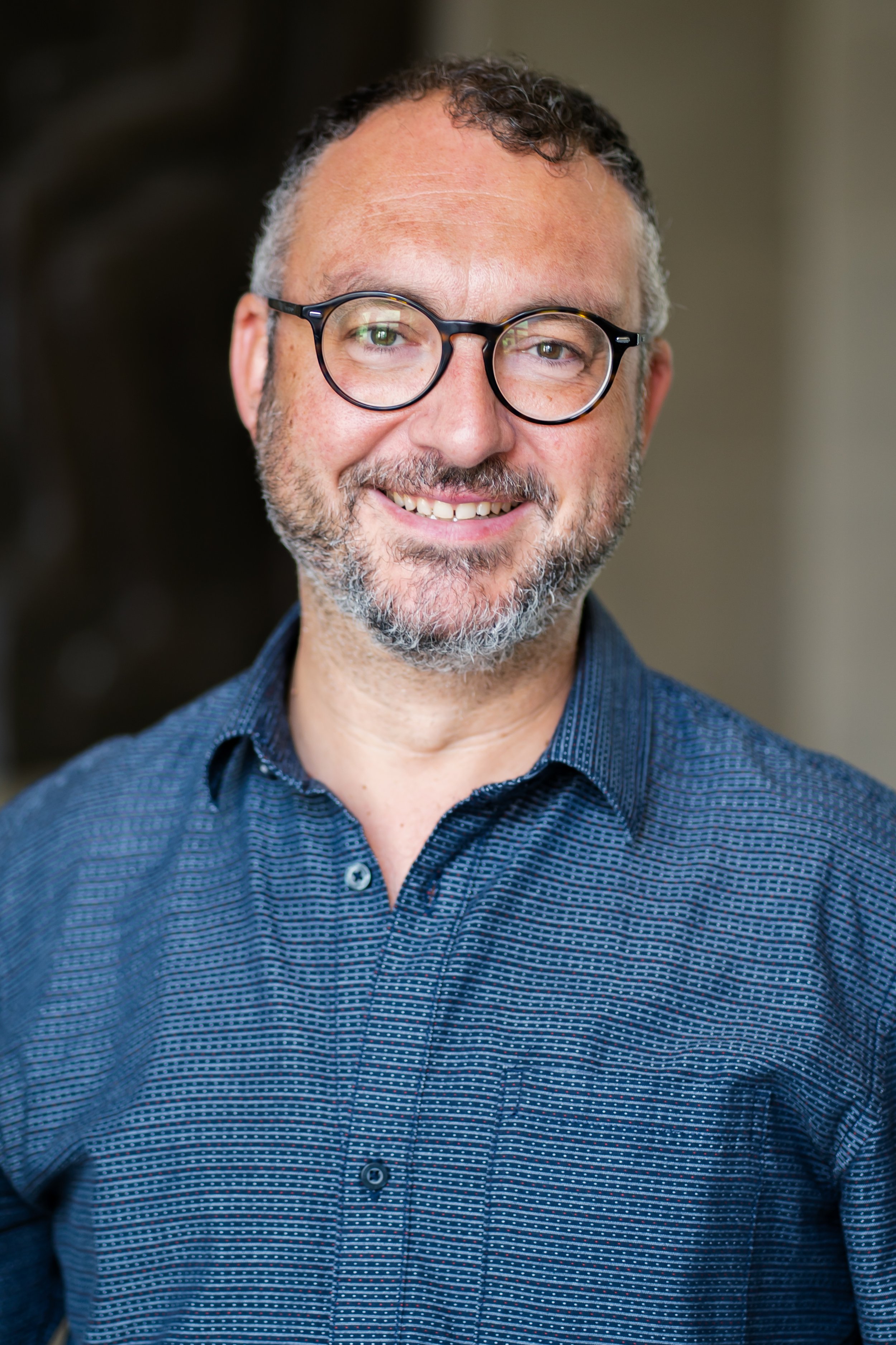 Fotografie eines lächelnden Mannes mit Brille, Bart und grau-braunen Haaren, trägt eine dunkelblaue, gemusterte Hemd, vor einem unscharfen, dunklen Hintergrund.