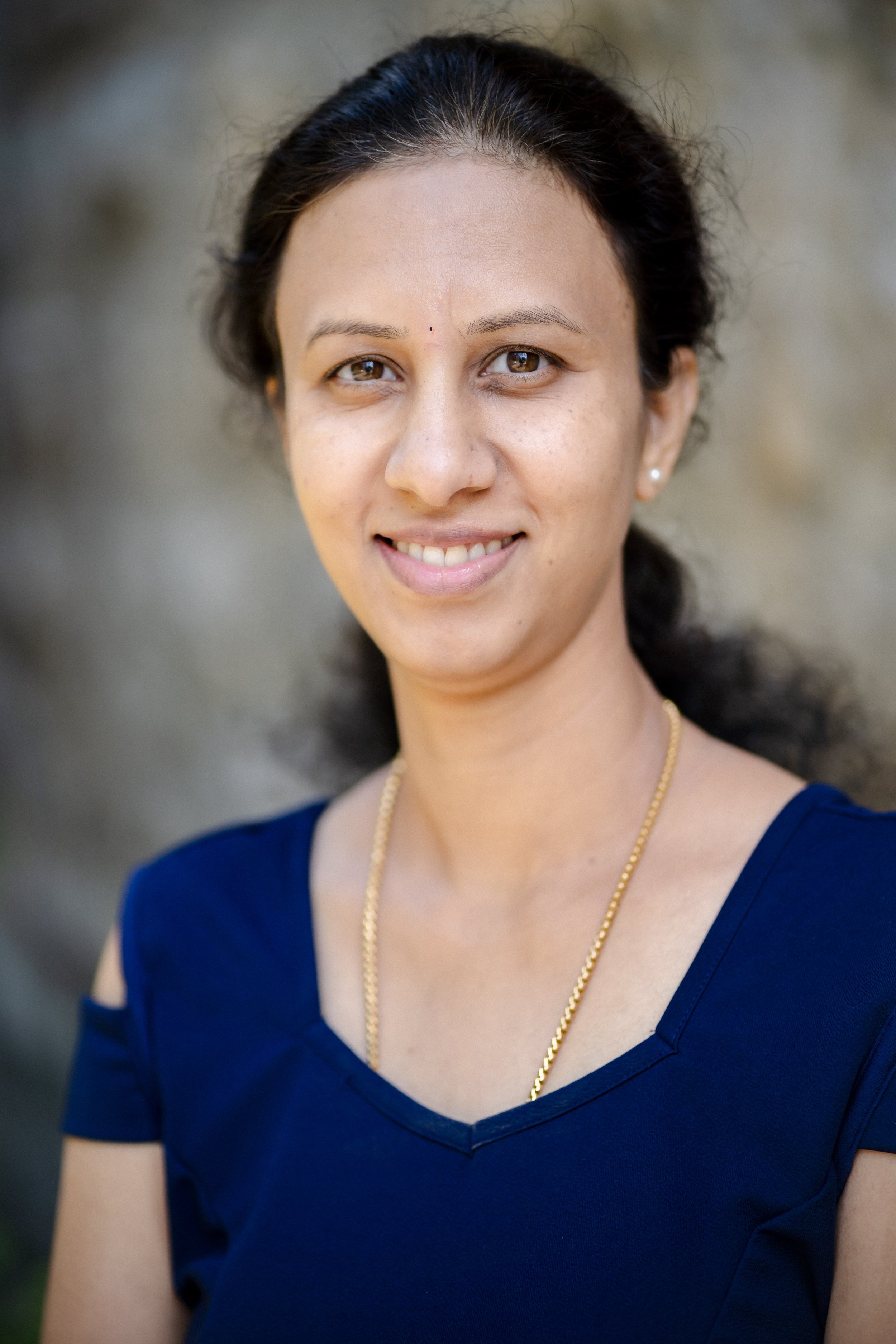 Nahaufnahme einer lächelnden Frau mit dunklen Haaren, die einen blauen V-Ausschnitt-Top trägt und vor einem unscharfen, neutralen Hintergrund steht.