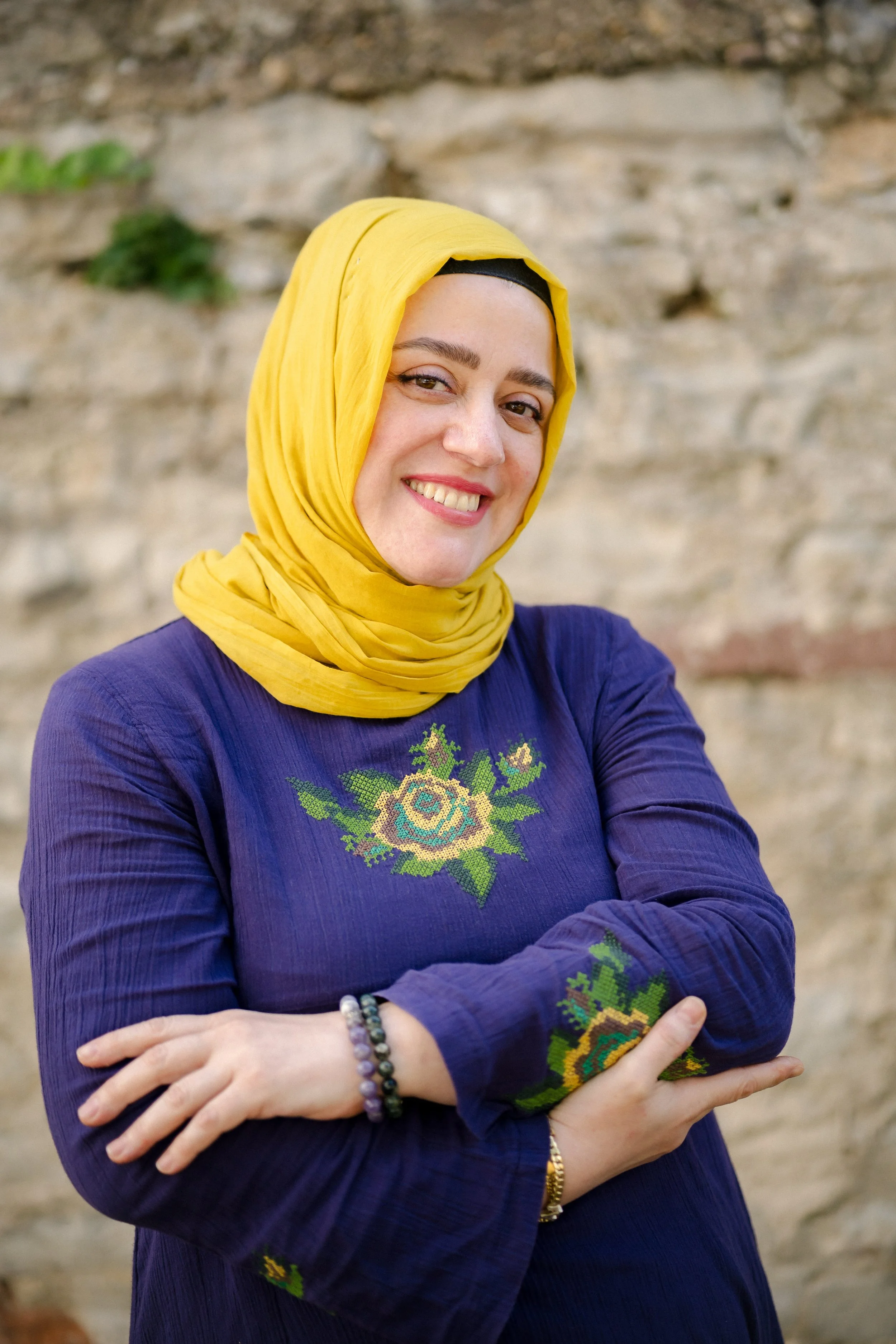 Eine lachende Frau mit gelbem Schal und blauer Bluse vor einer Steinmauer.