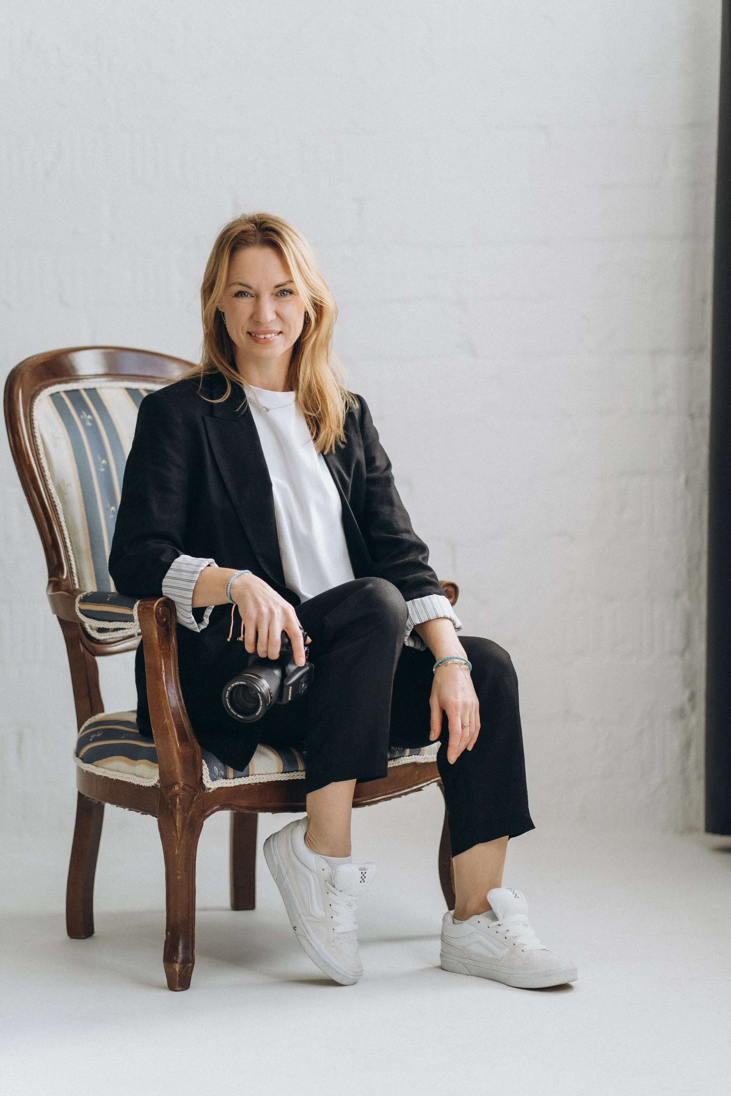 Eine junge Frau sitzt entspannt auf einem Vintage-Stuhl, trägt schwarze Kleidung, weiße Turnschuhe und hält eine Kamera in der Hand.