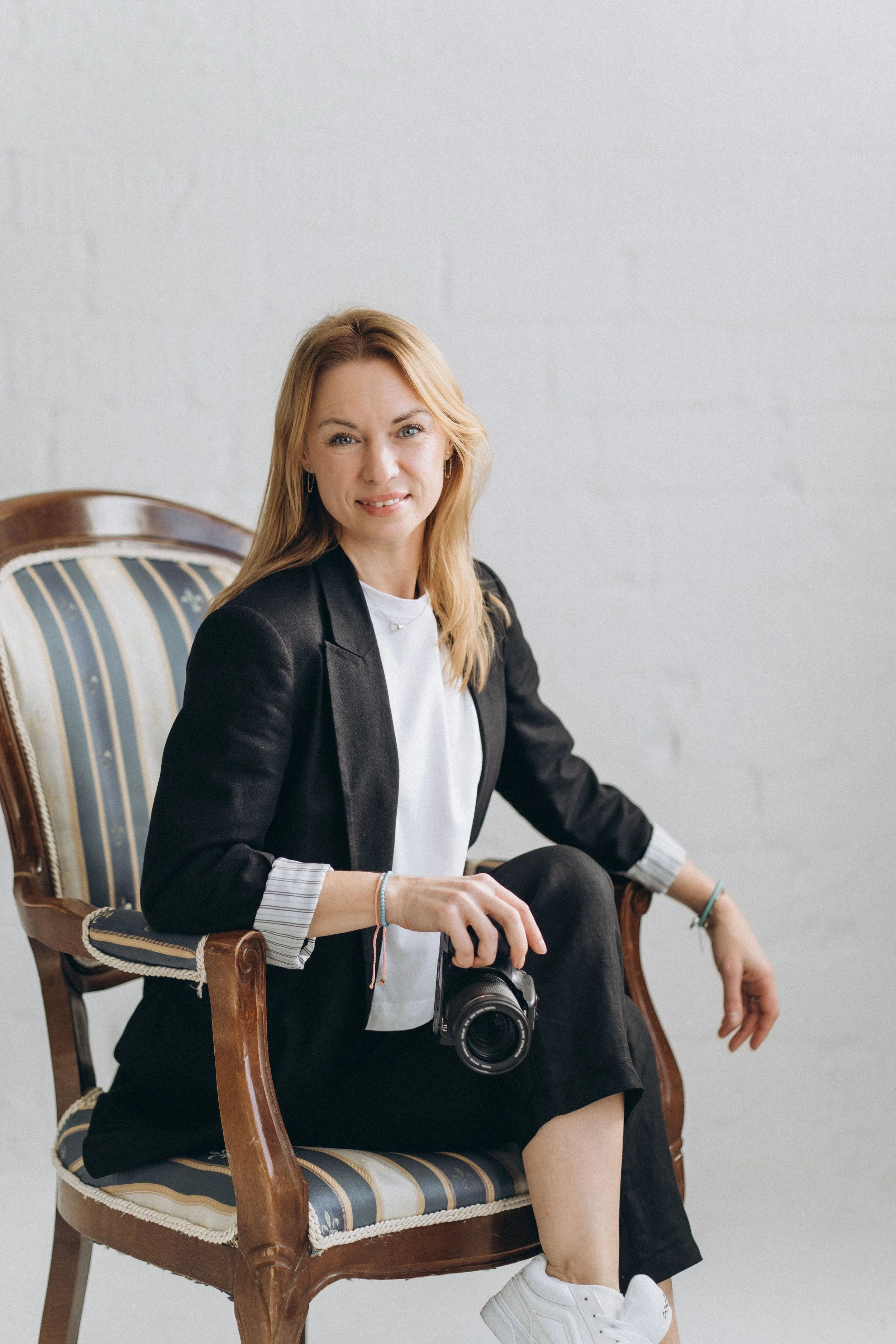 Eine Frau sitzt auf einem antiken Holzstuhl mit gepolstertem Sitz, trägt eine schwarze Jacke, weiße Schuhe und hält eine Kamera in der Hand, vor einer weißen Wand.