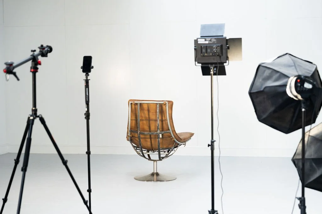 Photography studio setup with a vintage leather chair, multiple studio lights, two black umbrella lights, and a white wall background.