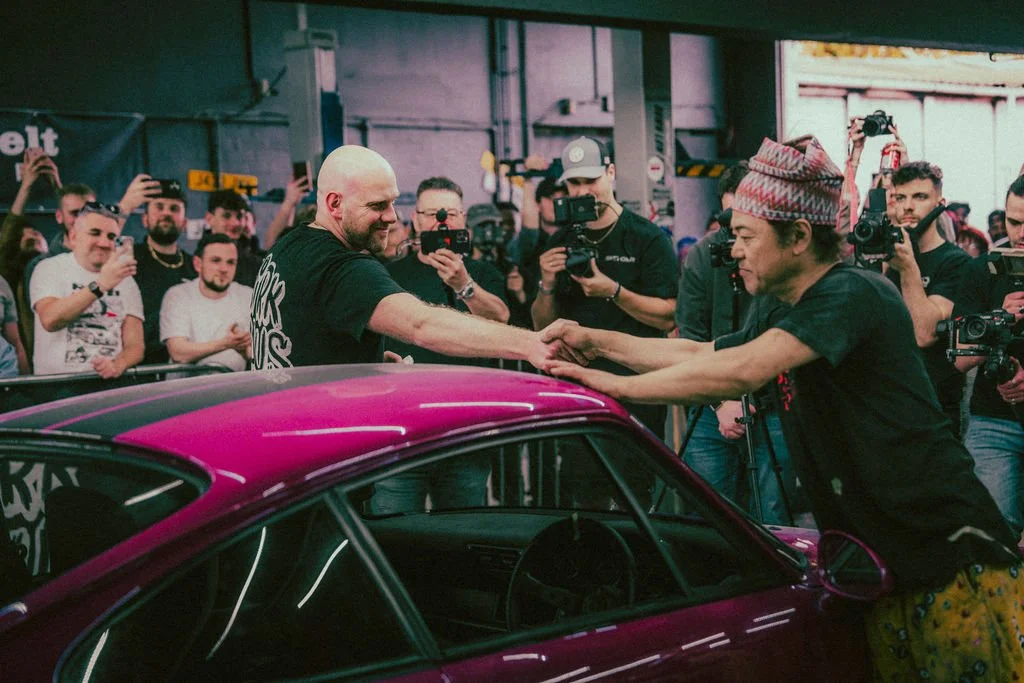 Two men are shaking hands across a pink car, while surrounded by a crowd of onlookers and photographers indoors.