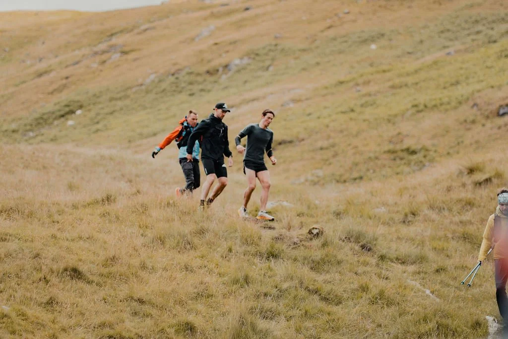 Three runners making their way down a grassy hillside with another hiker on the right, all dressed in outdoor gear.