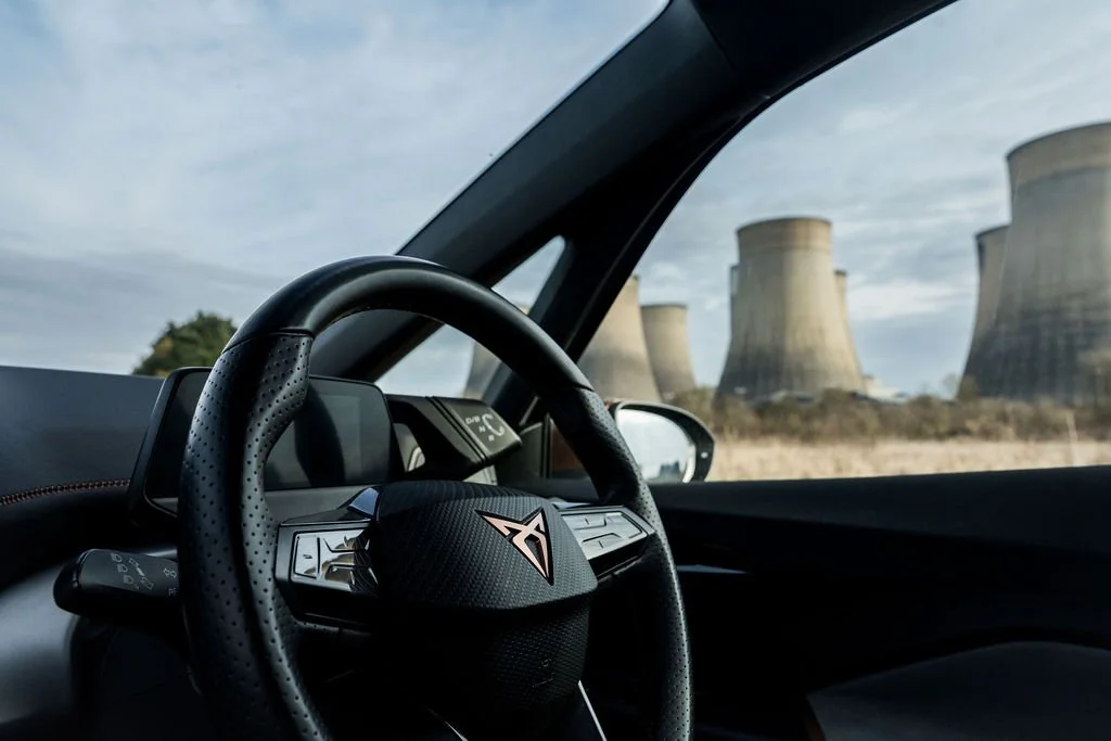 The interior of a sports car with a dashboard and steering wheel visible, showing cooling towers in the background through the windshield.