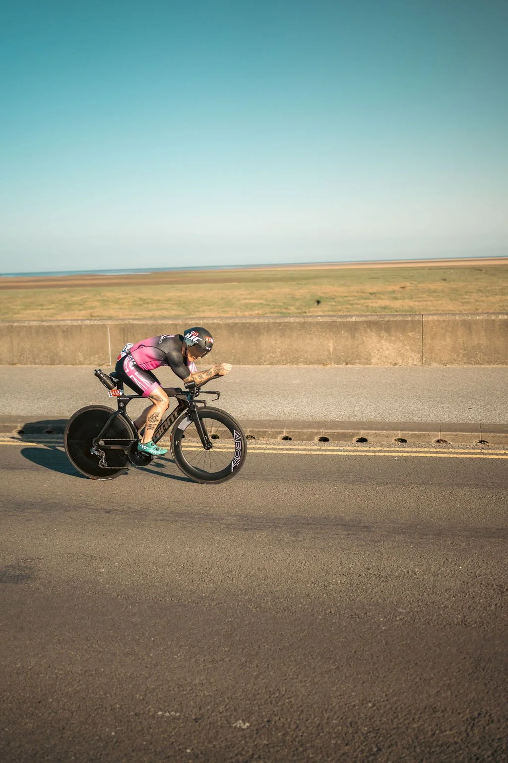 A cyclist wearing a helmet and racing gear riding a black time trial bike on a road near a beach or ocean, with a clear blue sky overhead.