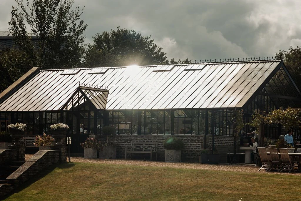 Greenhouse with glass walls and metal roof, surrounded by outdoor seating and greenery.