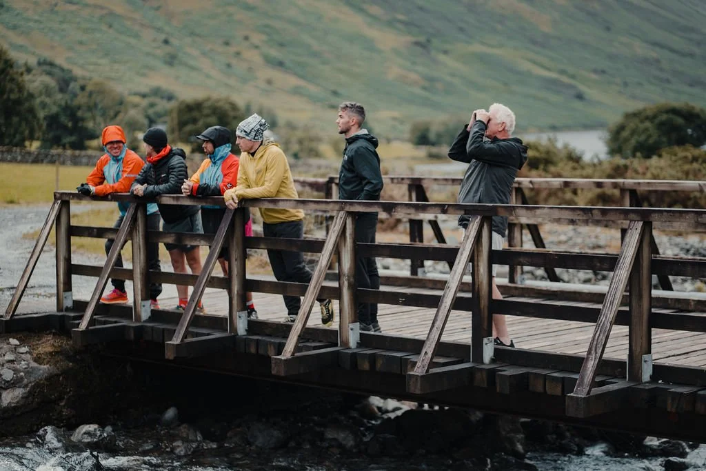 Group of six people on a wooden bridge overlooking a river with green hills in the background, some taking photos and others looking at the scenery.