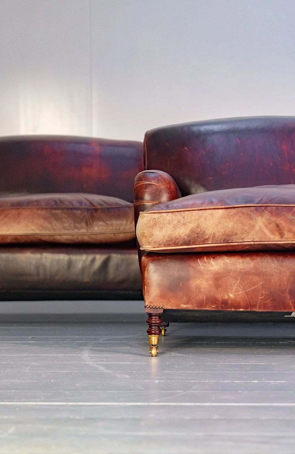 Close-up of two vintage leather armchairs with wooden legs on a gray wooden floor.