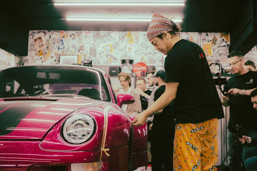 Akira Nakai with a patterned hat and bright yellow pants with colorful designs is working on a pink sports car in a crowded indoor space, with several people watching and some filming.