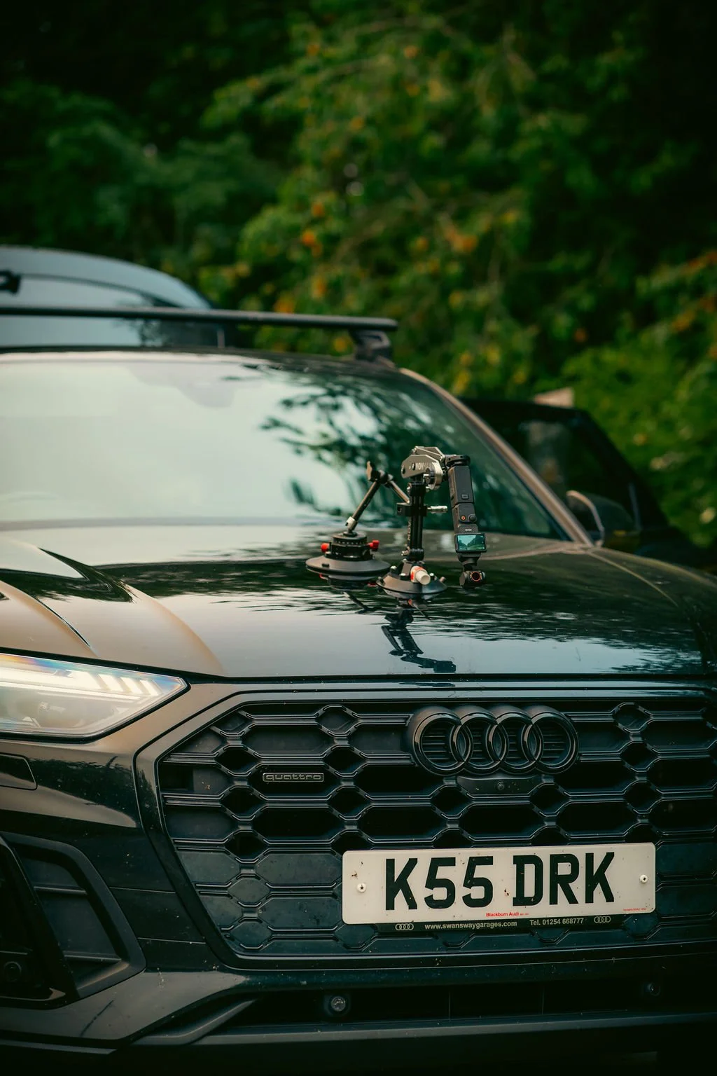 A black Audi car with a roof-mounted camera and stabilizer rig on the hood, parked outdoors with greenery in the background.