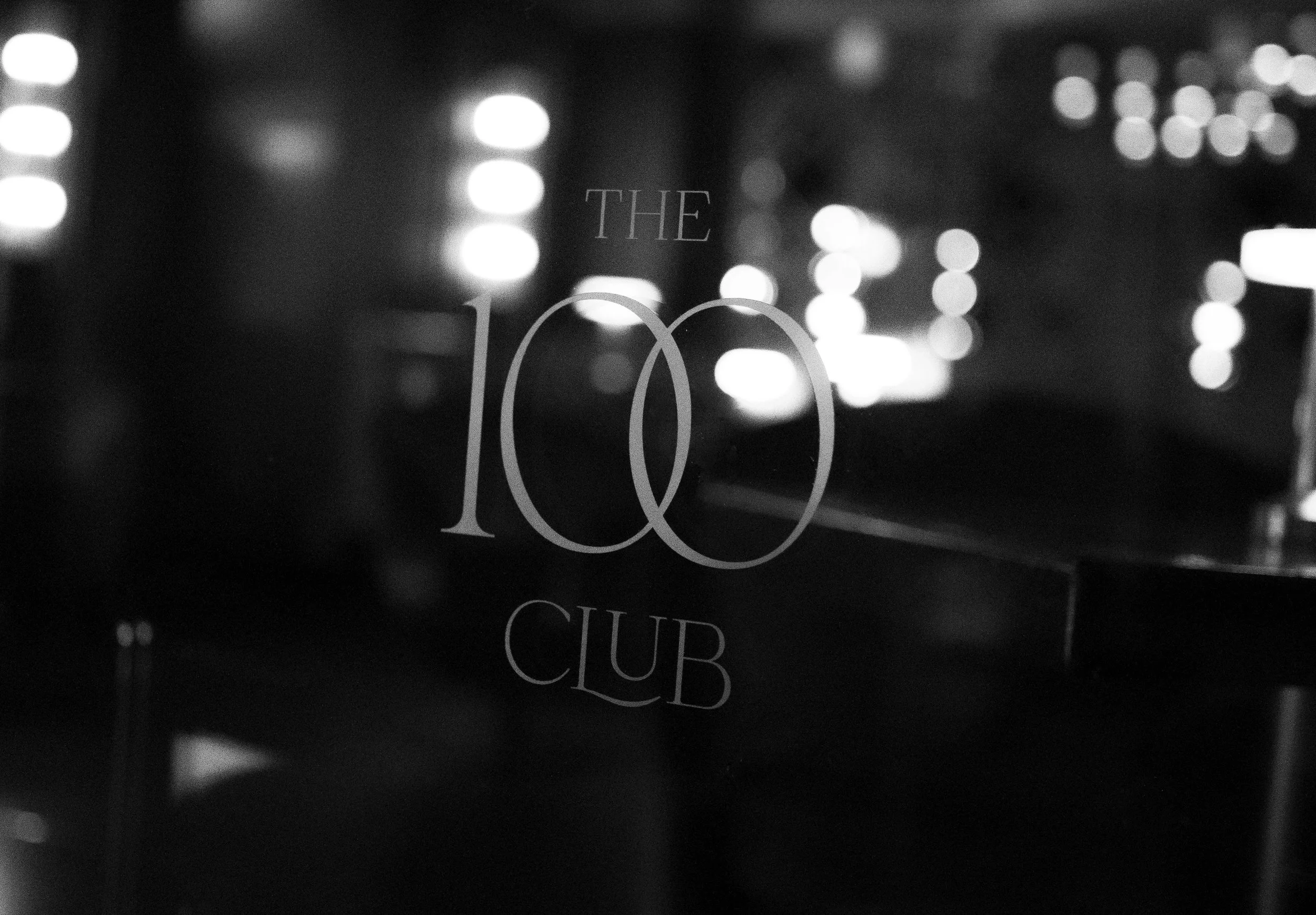 A black and white image showing the text 'The 100 Club' on a reflective surface with blurred city lights in the background.