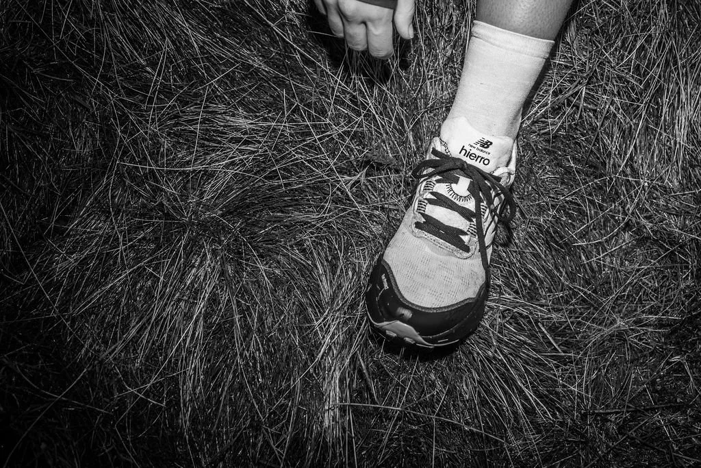 Close-up of a person's foot wearing a running shoe on grassy ground, with their hand holding the ankle.