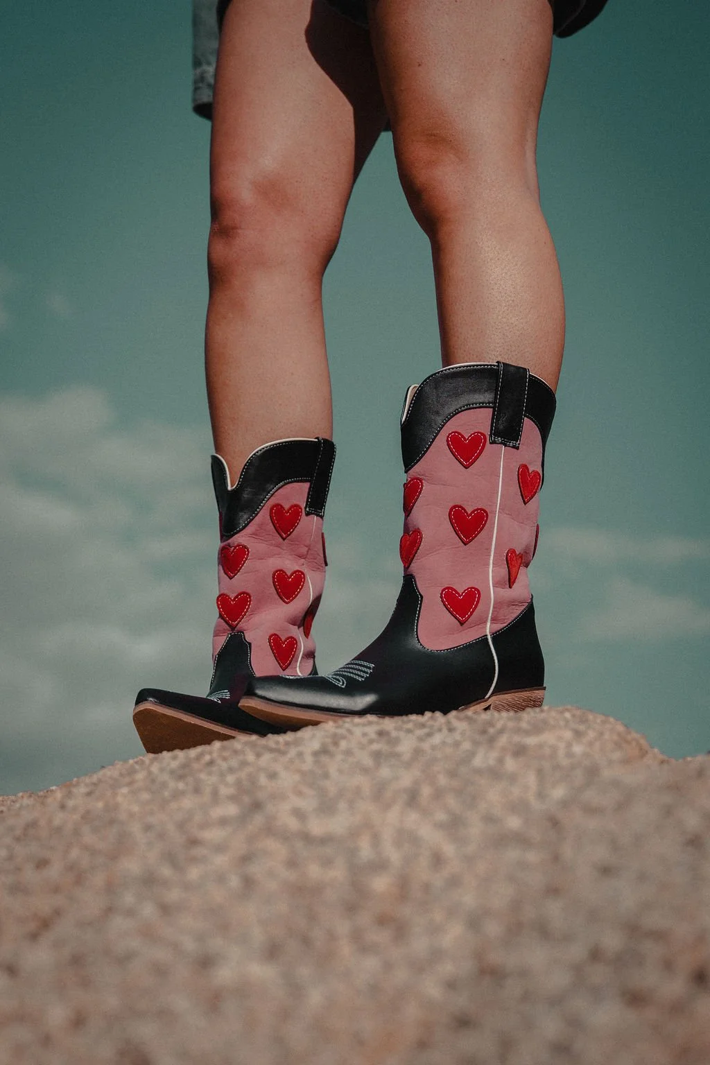 Person standing on sand wearing cowboy boots decorated with red hearts, under a cloudy sky.