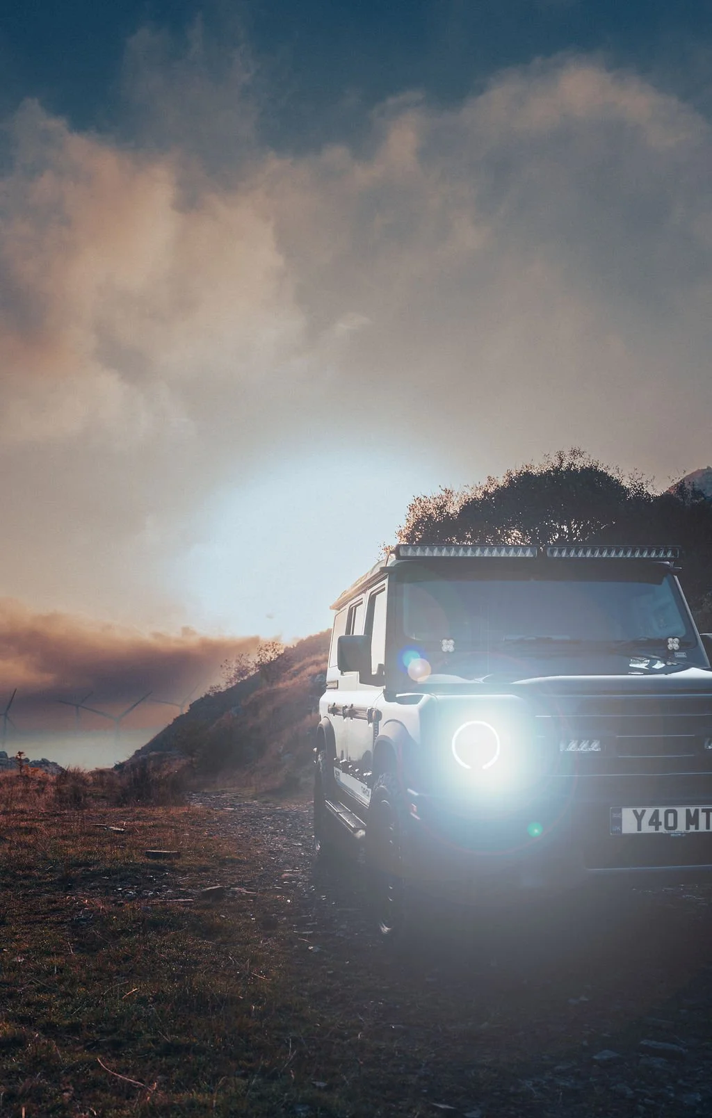 A dark-colored SUV parked on a dirt trail with the sun setting behind a hillside, clouds in the sky, and wind turbines in the distance.
