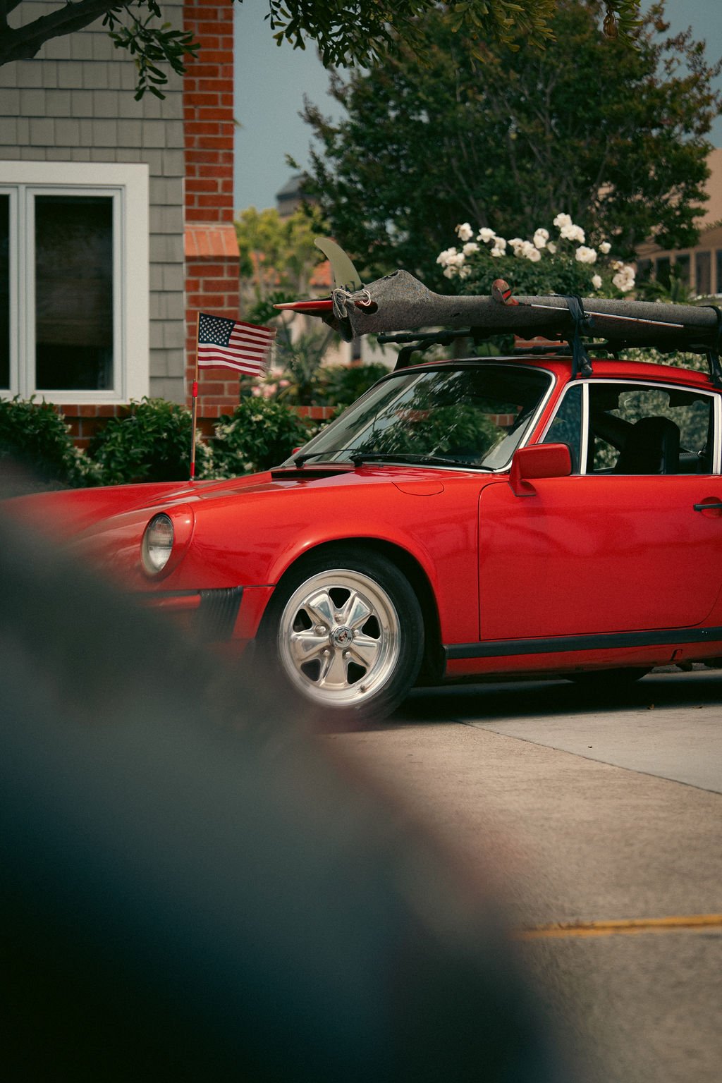 A red Porsche parked in a driveway with a surfboard on top, secured with a strap. An American flag on a small pole is attached to the car, and there is a house with a window, bushes, and blooming white flowers in the background.