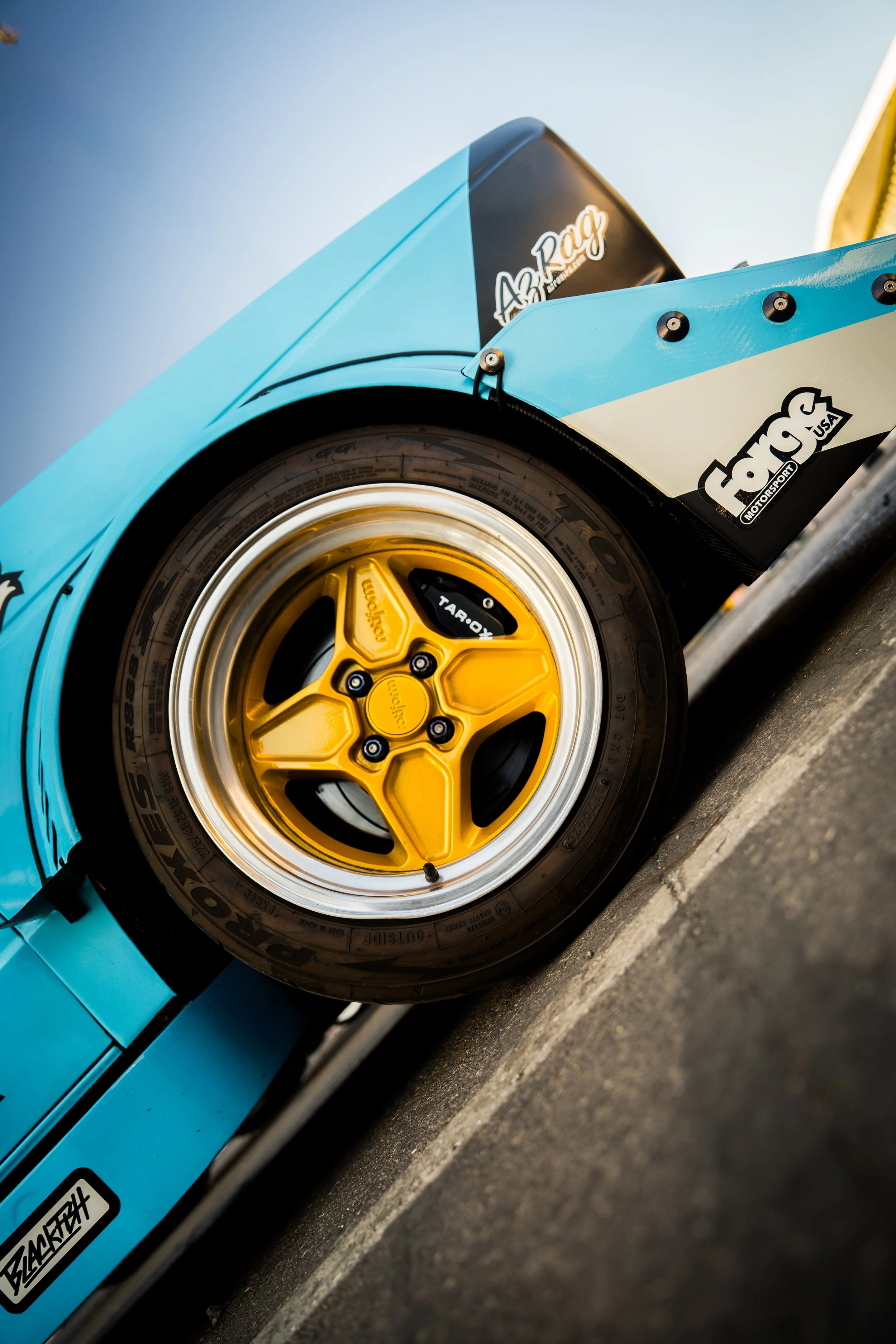 Close-up of a race car's front wheel with a yellow rim, black tires, and a light blue body with sponsor decals, including 'Frog' and 'Asphalt.'