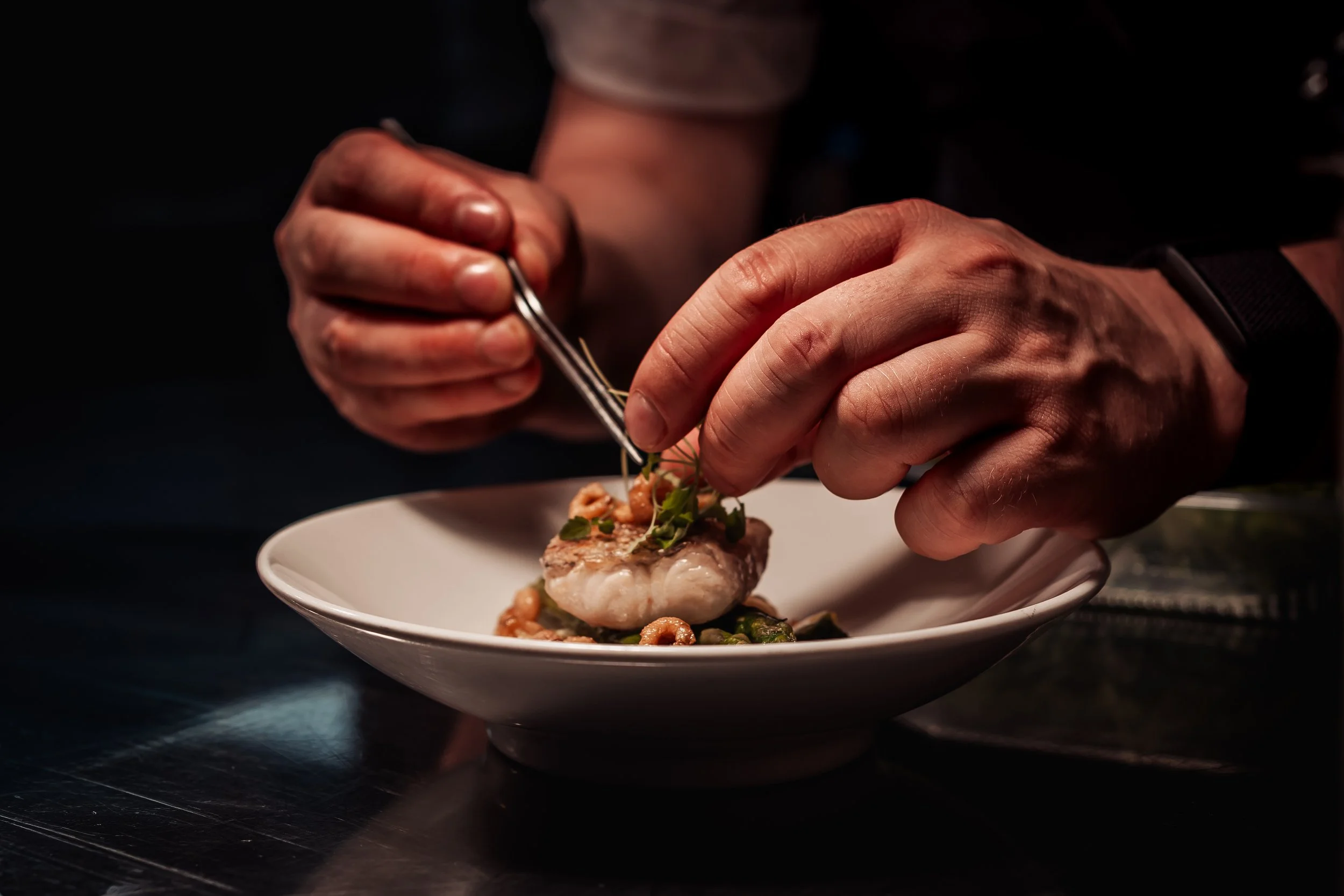 Chef garnishing a plated dish with herbs using tweezers in a dark kitchen setting.